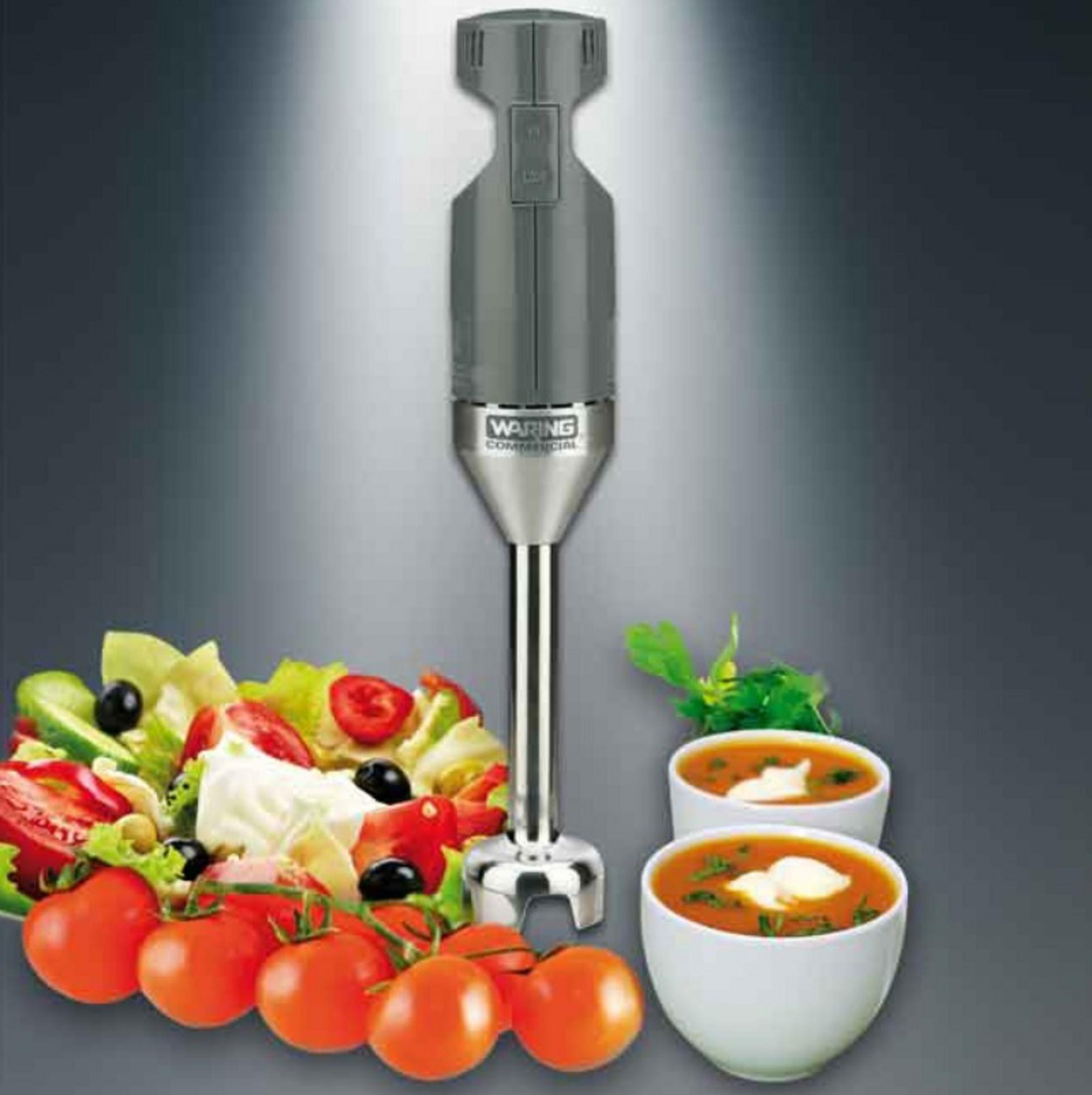Un frullatore ad immersione grigio vicino a insalata fresca, pomodori e due ciotole di zuppa su sfondo grigio.