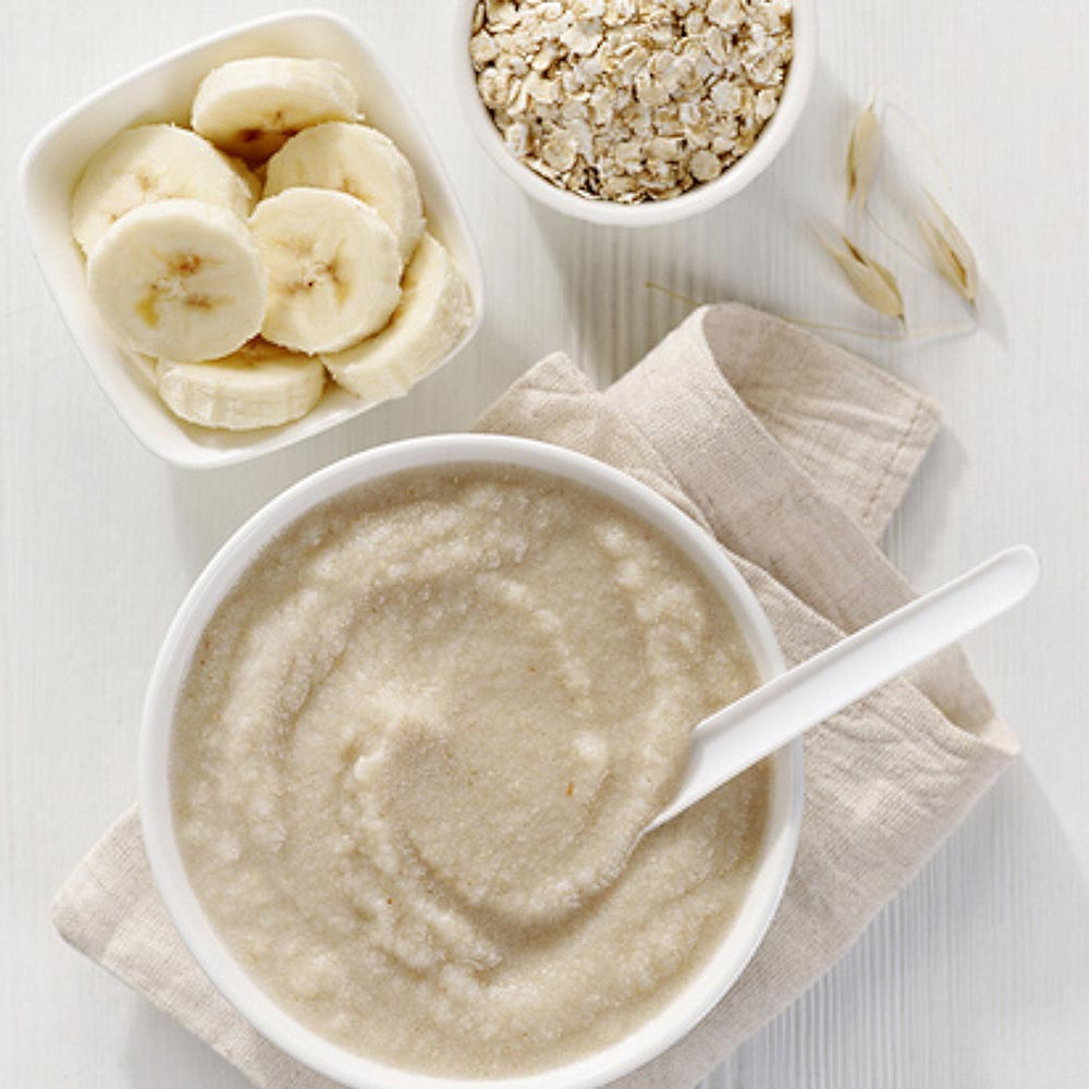 Una ciotola di porridge con cucchiaio, accompagnata da fette di banana e avena secca su fondo di legno bianco.