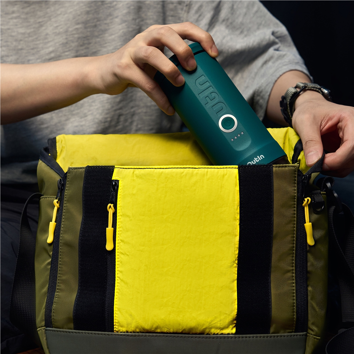 Una persona coloca una cafetera verde oscura en una bolsa verde oliva y amarilla.