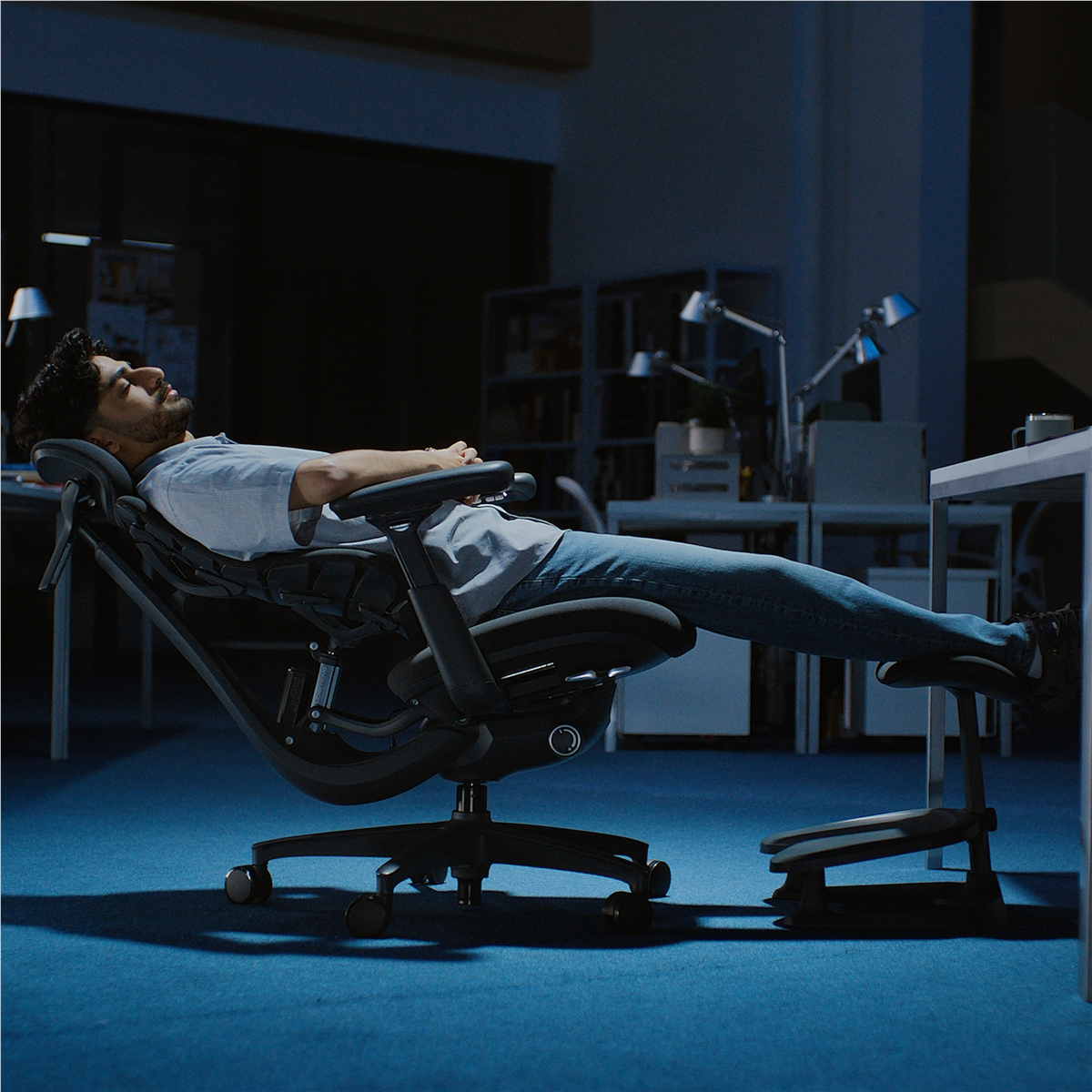 Hombre descansa en una silla de oficina ergonómica con las piernas en un reposapiés en una oficina oscura.