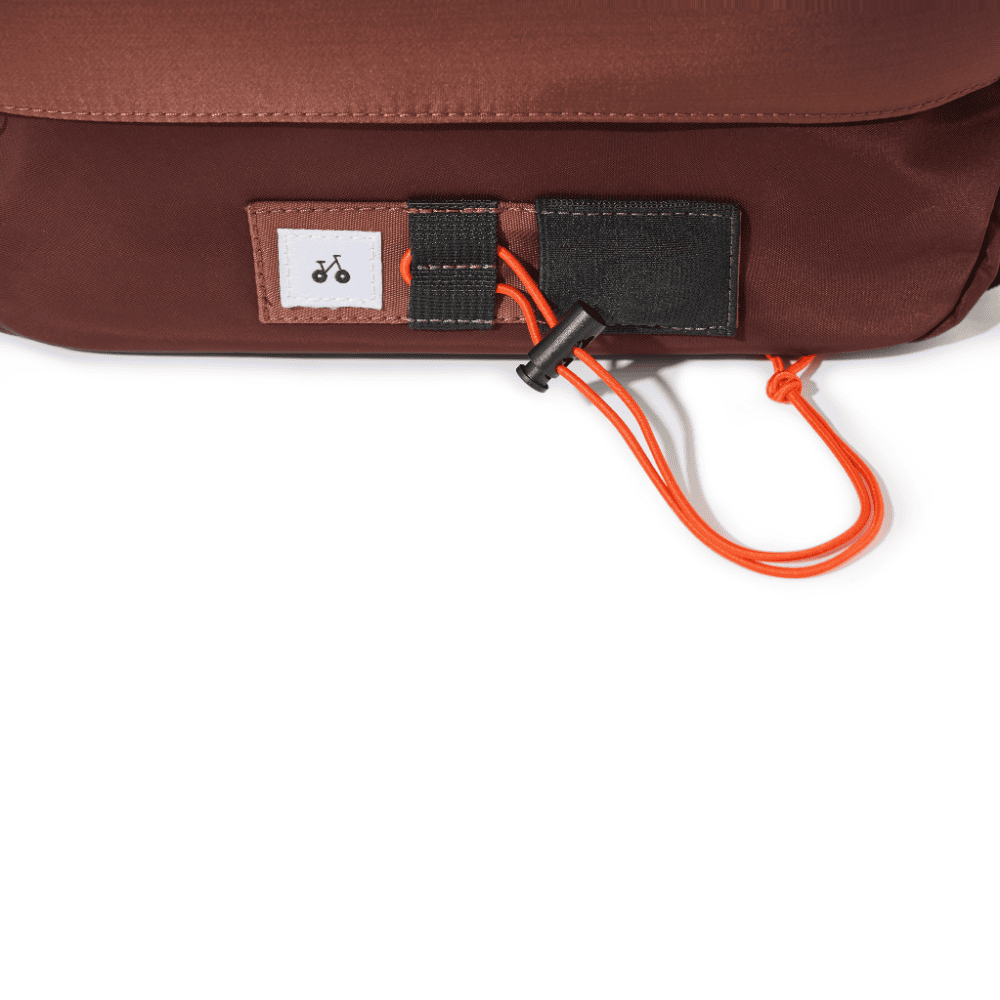 Un bolso marrón tiene una etiqueta blanca con un icono de bicicleta y un cordón naranja con tope negro.