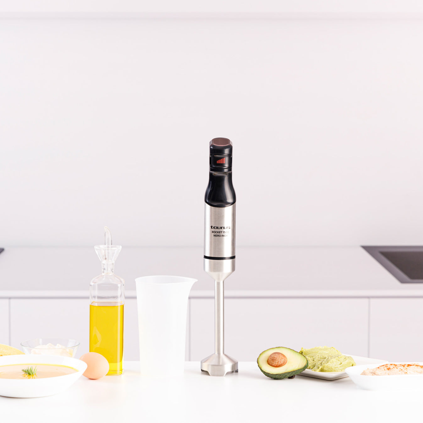 Un minipimer argentato si trova su un tavolo accanto a olio, un avocado e vari cibi.