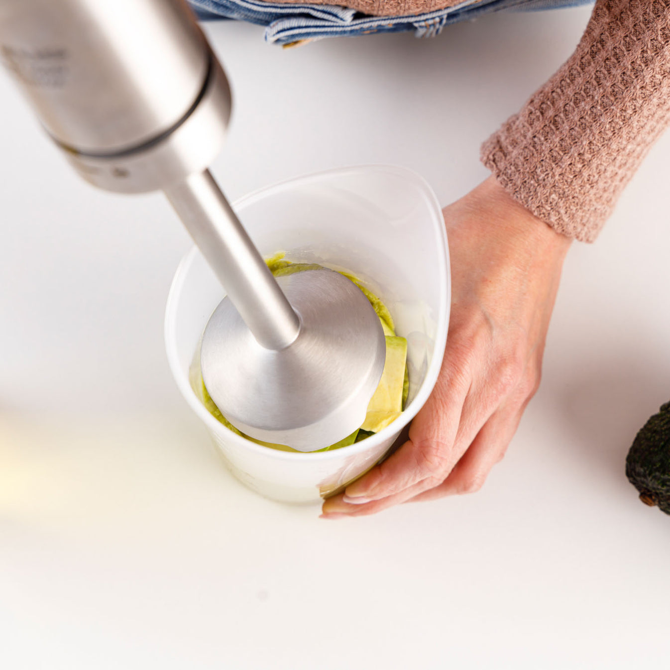 Una persona usa un minipimer in un bicchiere di plastica con pezzi di avocado.