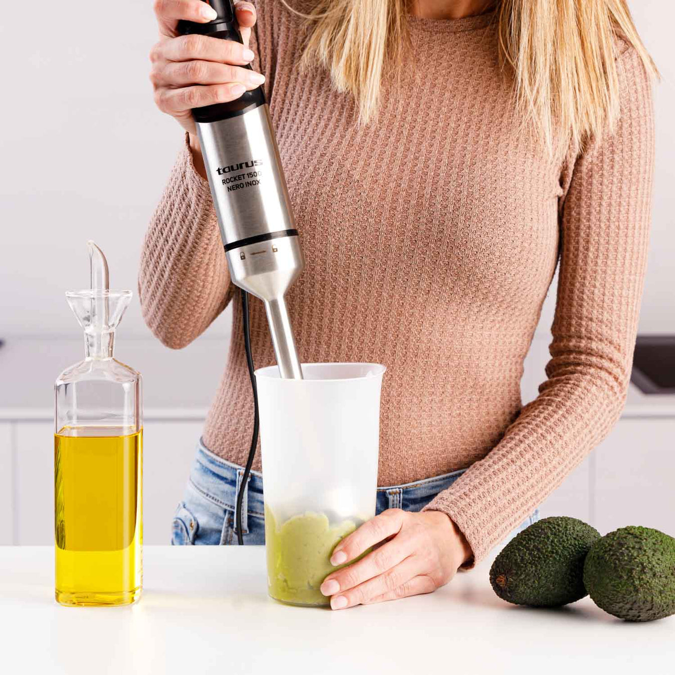 Una donna frulla pasta verde in un bicchiere con un minipimer, accanto a olio d'oliva e avocado.