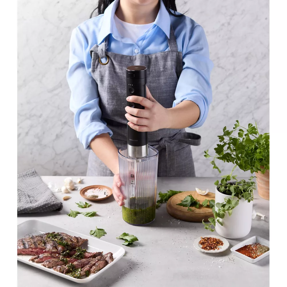 Una donna con camicia blu e grembiule grigio frulla erbe in un bicchiere accanto a carne e spezie su un tavolo.