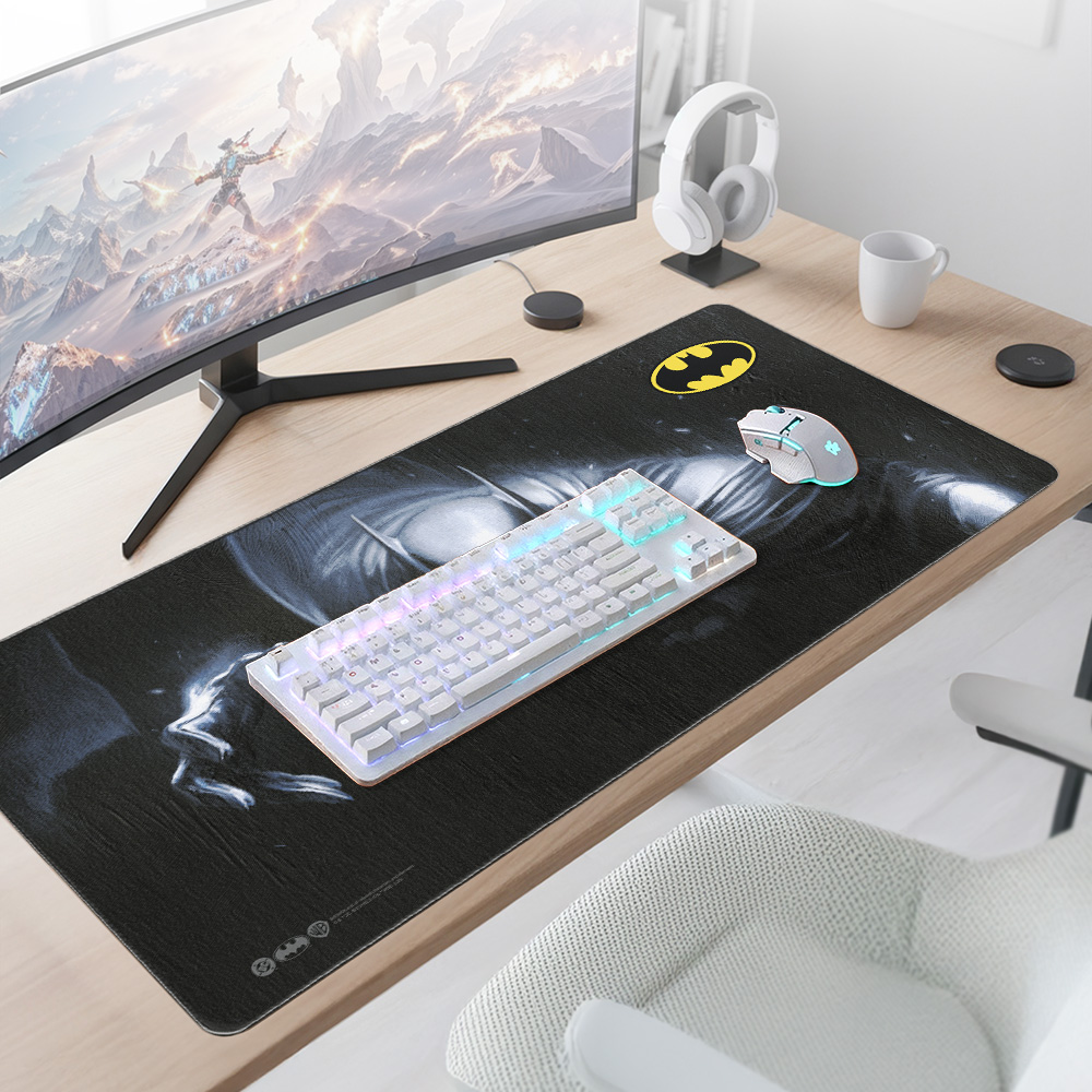 Una scrivania pulita con monitor curvo, tappetino Batman, tastiera bianca, mouse, cuffie e una tazza.