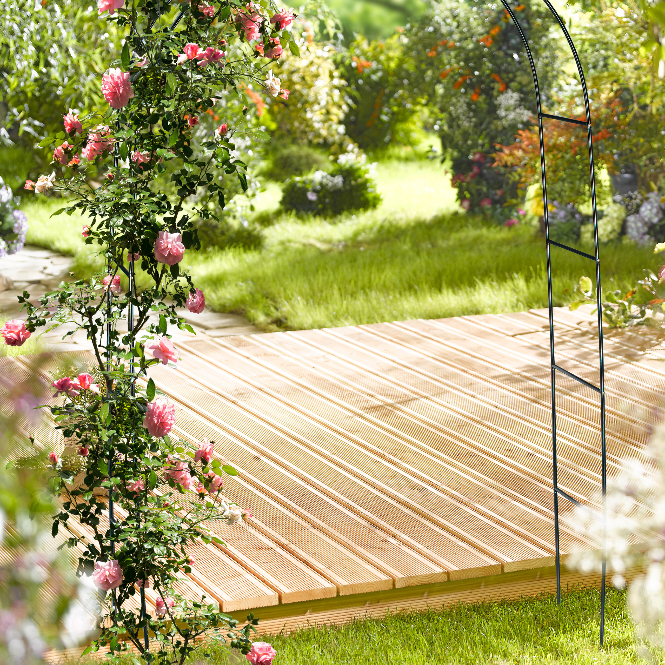 Ein hölzernes Deck im Garten, eingerahmt von einem schwarzen Metallbogen, der mit pinken Rosen bewachsen ist.