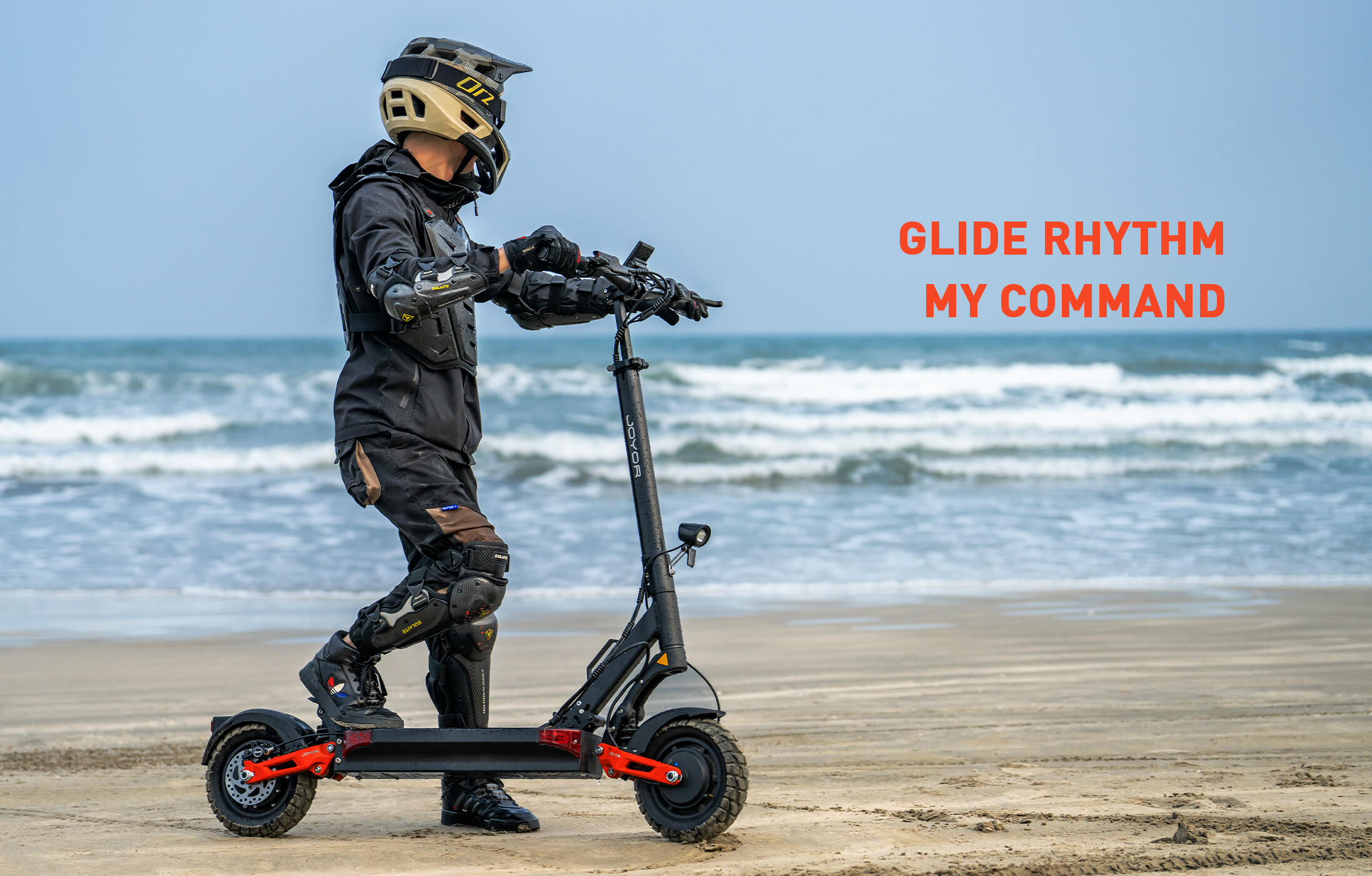 Un ciclista con equipaggiamento protettivo e casco si trova su un monopattino elettrico in spiaggia.