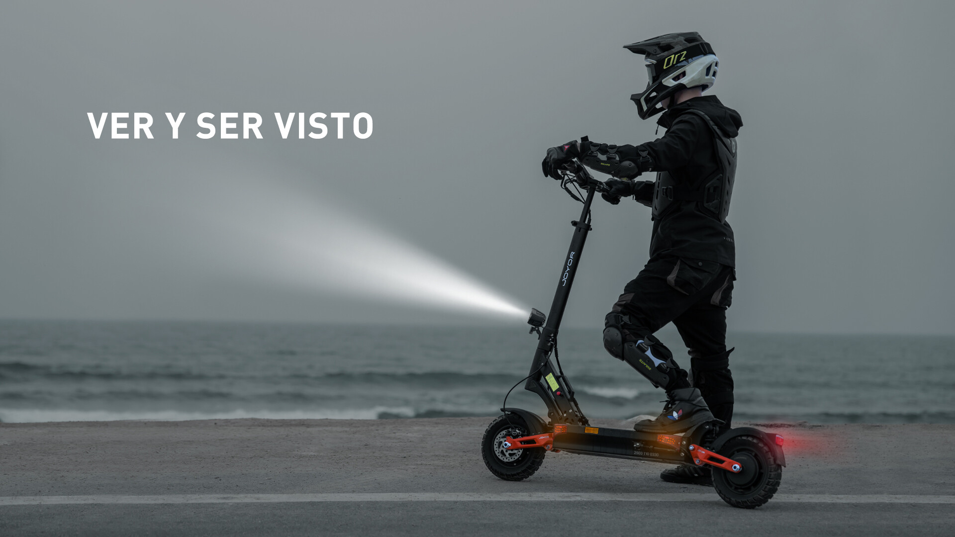 Un guidatore con casco su monopattino elettrico al mare, con faro acceso su una strada grigia.