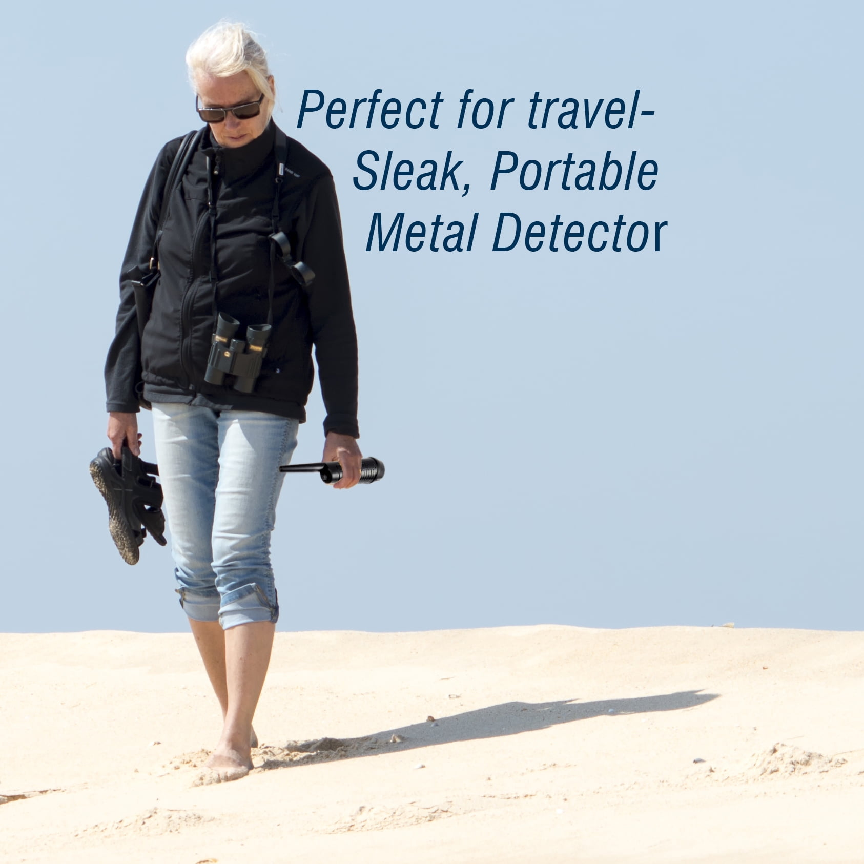 Frau mit Sonnenbrille und Fernglas geht im Sand und hält ein Gerät.