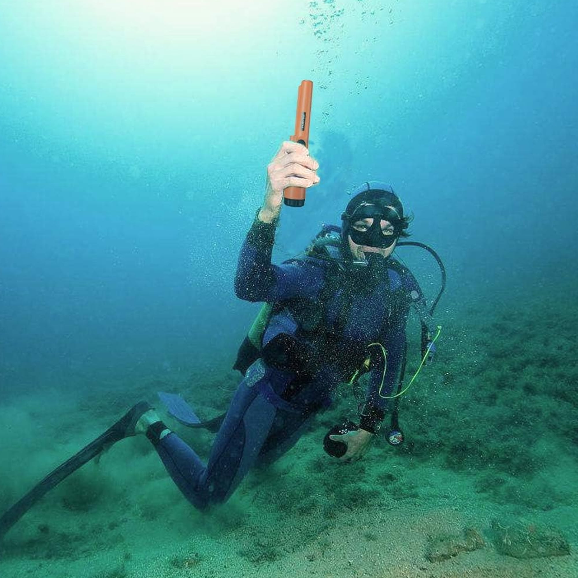 Ein Taucher in blauer Ausrüstung schwimmt im klaren Wasser über sandigem Grund und hält ein orangefarbenes Gerät hoch.