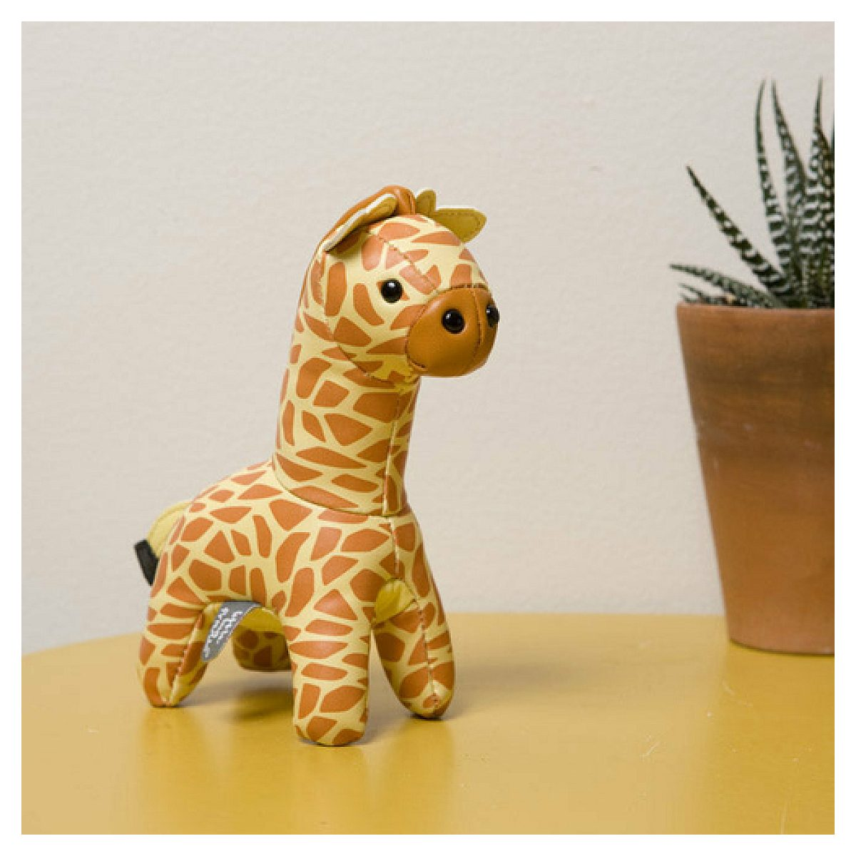 Eine kleine Giraffe aus Lederimitat steht auf einem gelben Tisch. Rechts steht eine Topfpflanze.