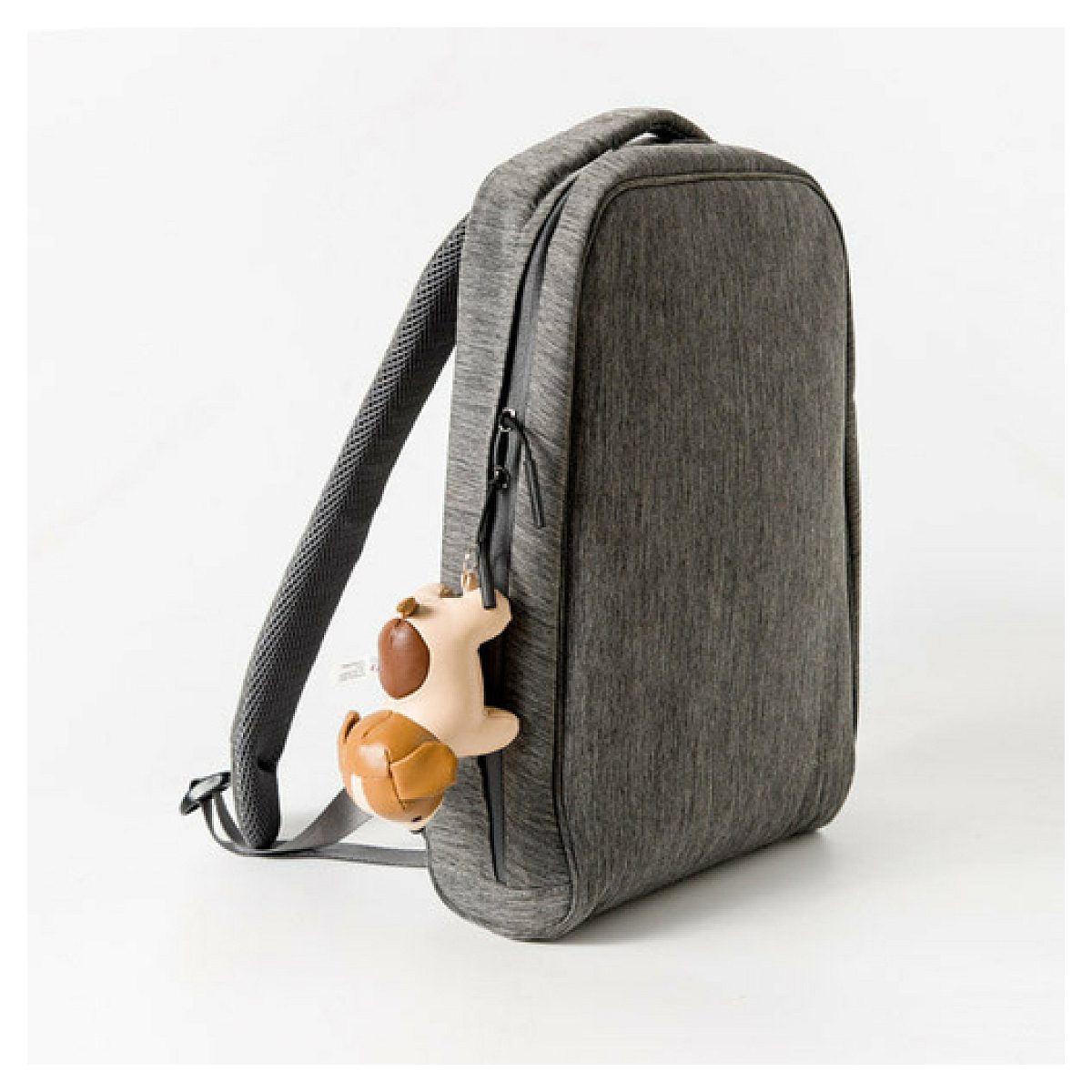 Ein grauer Rucksack mit einem kleinen braun-weißen Hundeanhänger am Reißverschluss vor weißem Hintergrund.