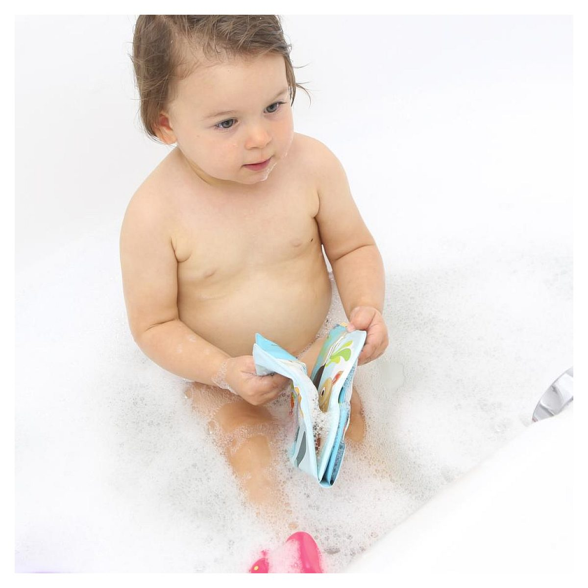 Ein Kleinkind sitzt in einer weißer Badewanne voller Schaum und hält ein kleines blaues Badebuch.