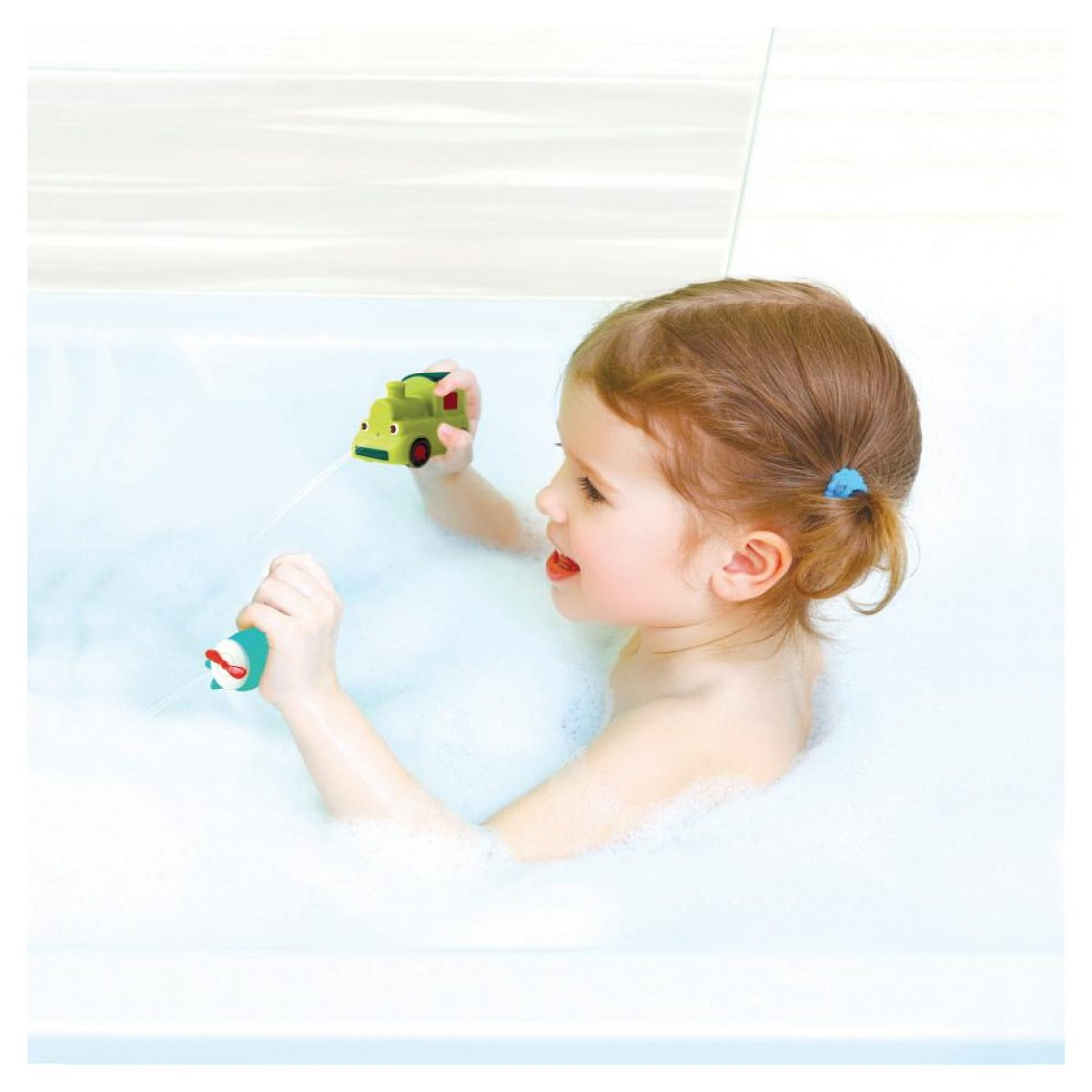 Kleines Mädchen sitzt in einer Badewanne voll Schaum und spielt mit zwei bunten Wasserspielzeugen.