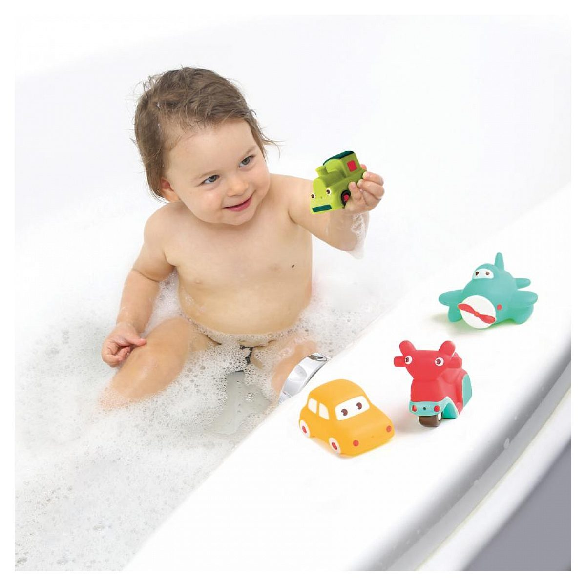 Ein Kleinkind spielt mit einem grünen Spielzeugzug in einer weißen Badewanne voller Schaum, neben drei weiteren Fahrzeugen.