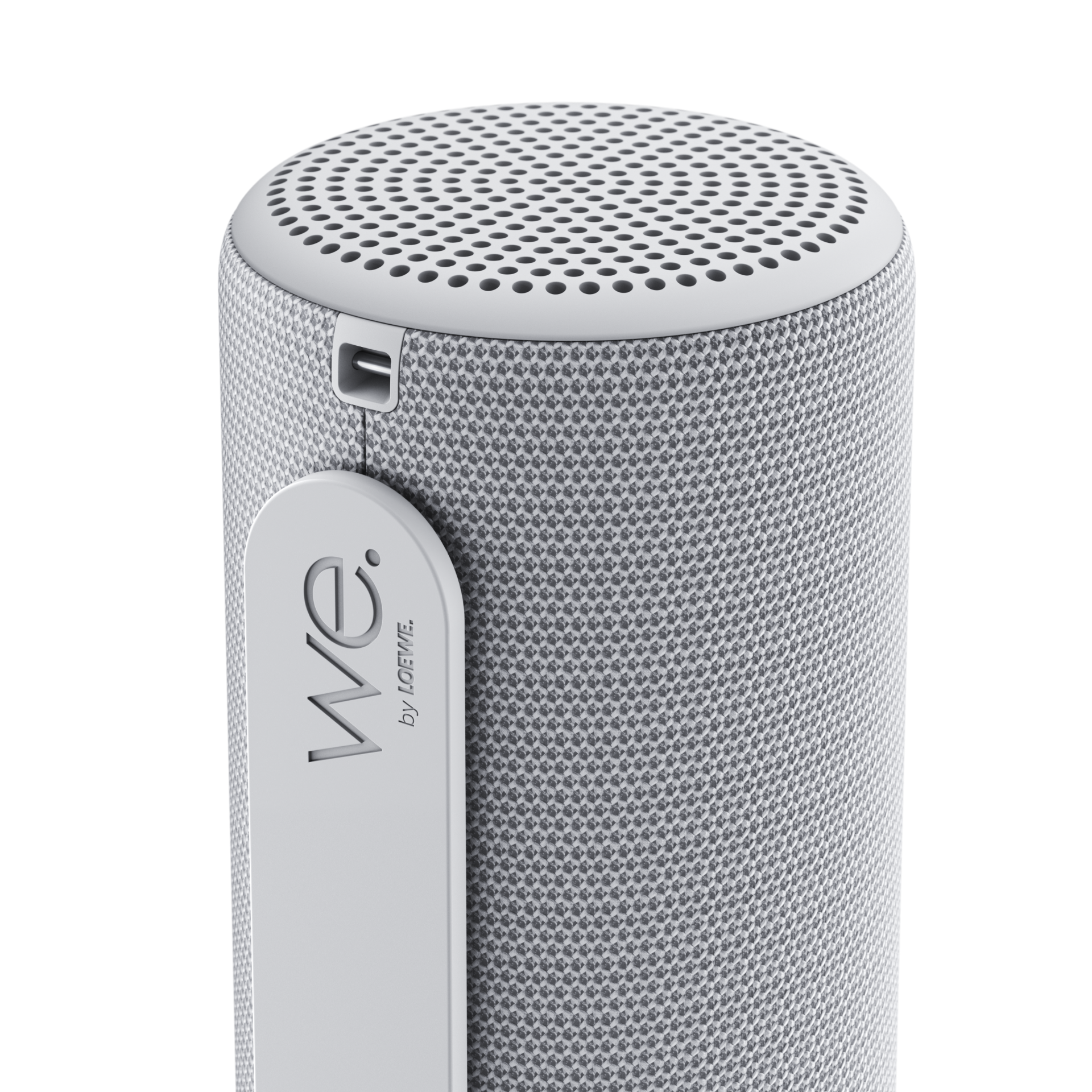Ein hellgrauer, zylindrischer Bluetooth-Lautsprecher mit Stoffbezug und einer Lasche mit der Aufschrift We. by LOEWE.