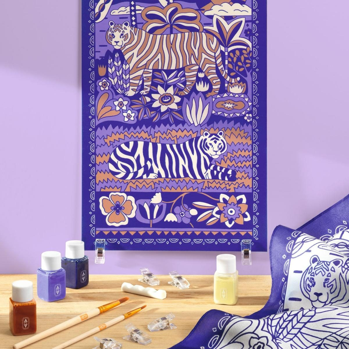 Ein lilafarbenes Poster mit Tigern in Dschungelszene, auf Holztisch mit Farbdosen und Pinseln platziert.
