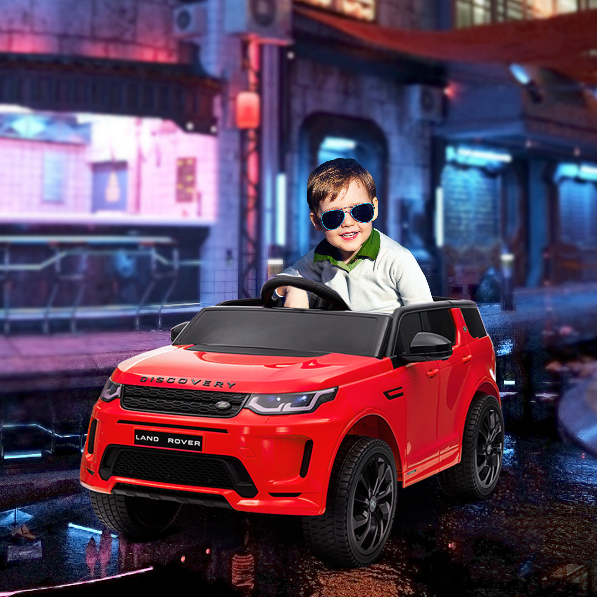 Un bambino con gli occhiali da sole guida una macchinina rossa Land Rover su una strada illuminata.