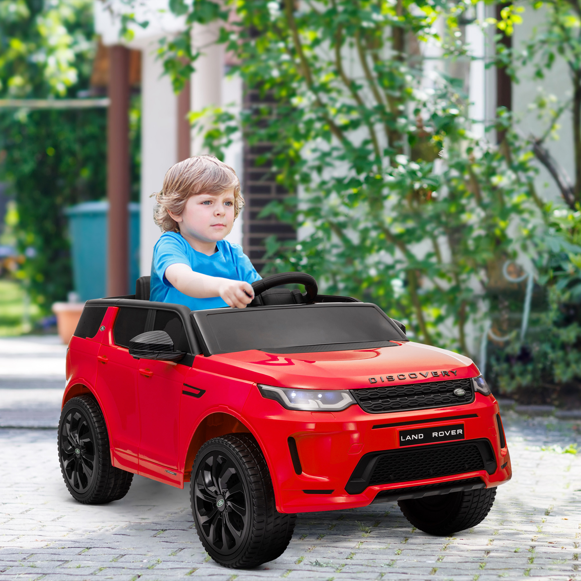 Un bambino biondo guida una macchinina rossa Land Rover all'aperto.