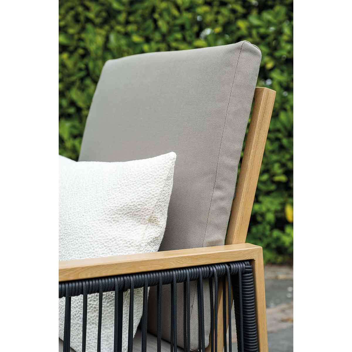 Wooden armchair with black rope, grey back cushion, and white throw pillow against green foliage.