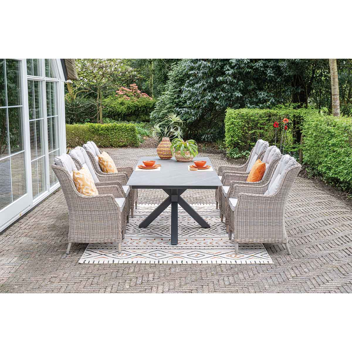 Wicker patio dining set with a table and chairs on a stone terrace in a garden.