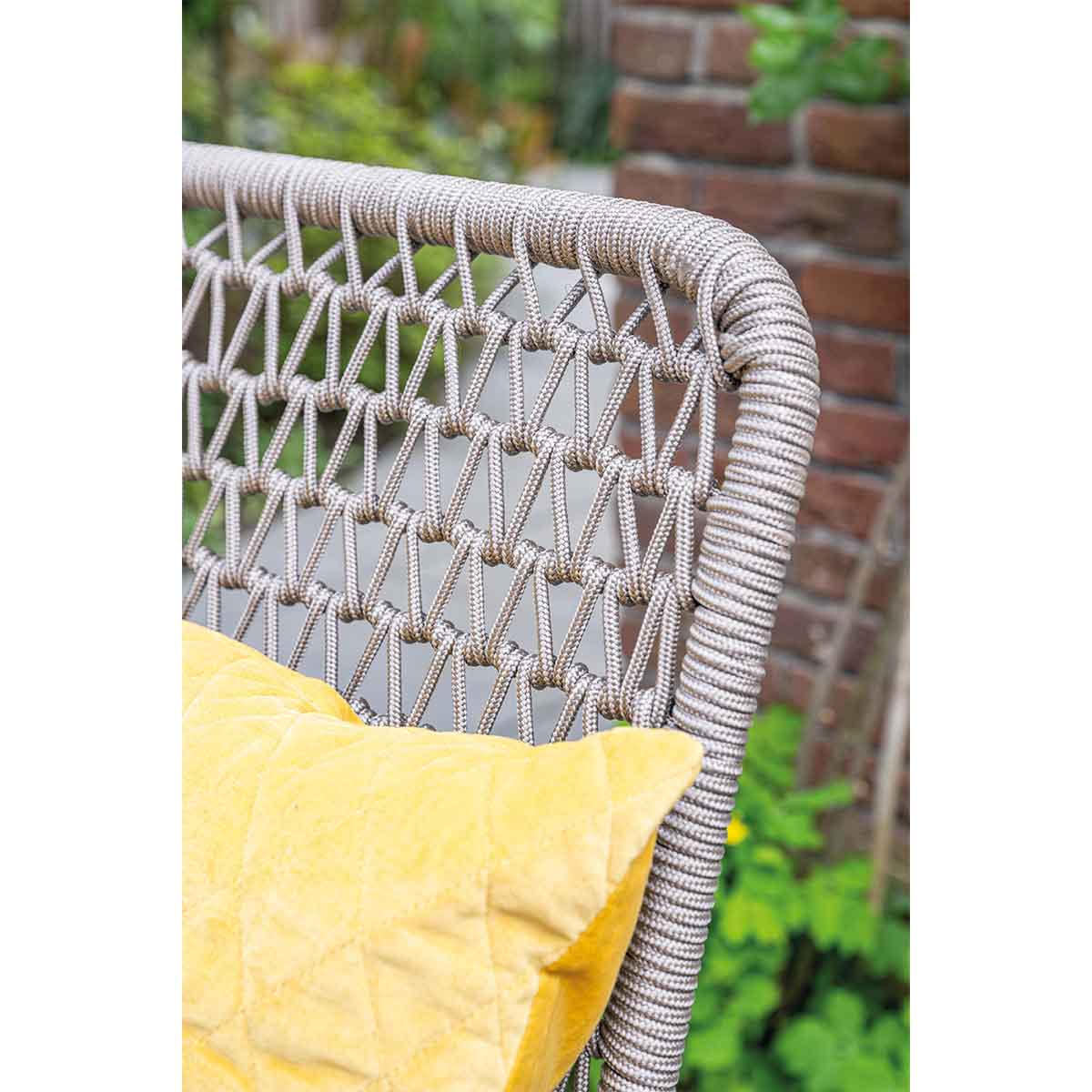 Close-up of a grey rope-woven chair with a yellow pillow in front of a brick wall.