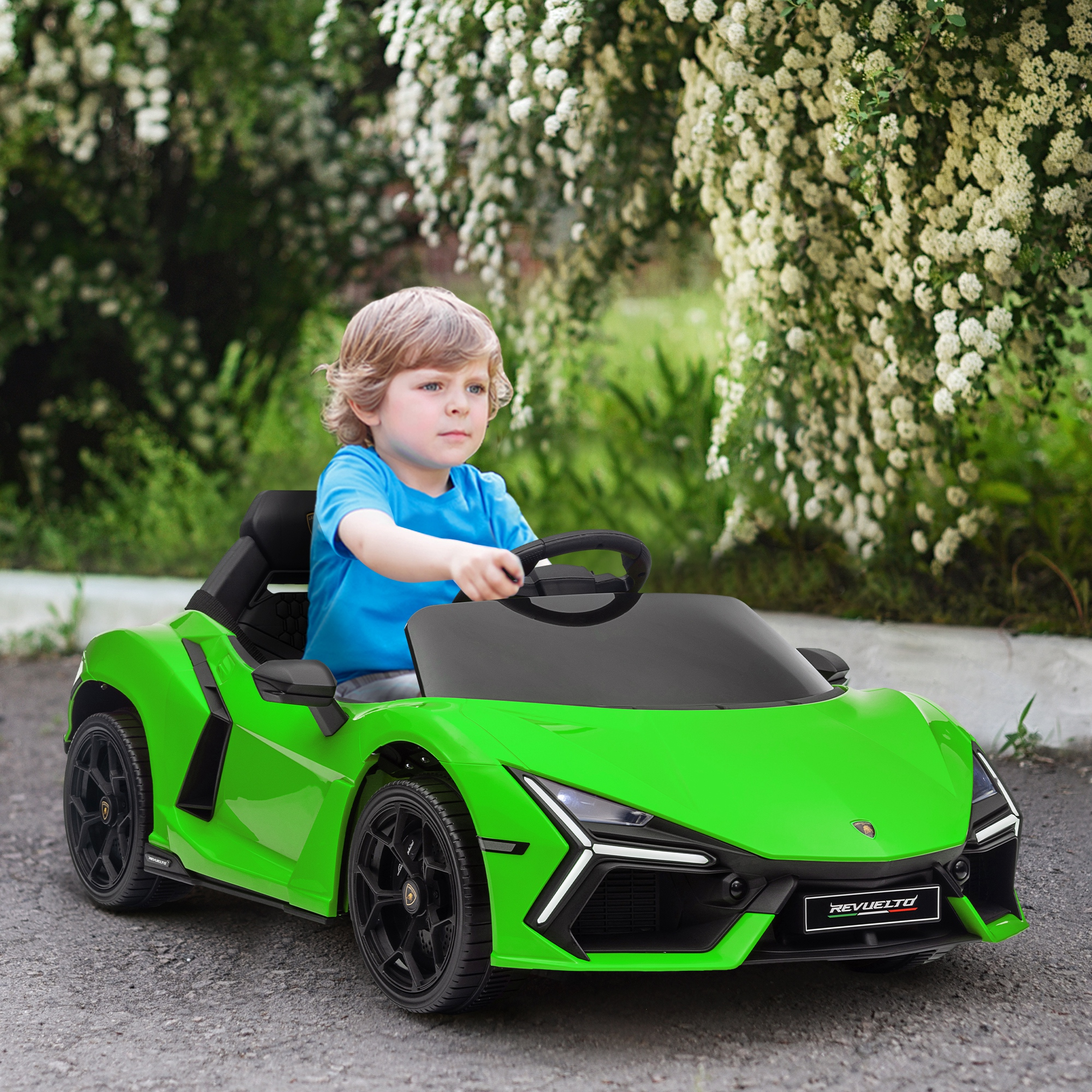 Un ragazzo con una maglietta blu siede su un'auto giocattolo verde davanti a cespugli fioriti.