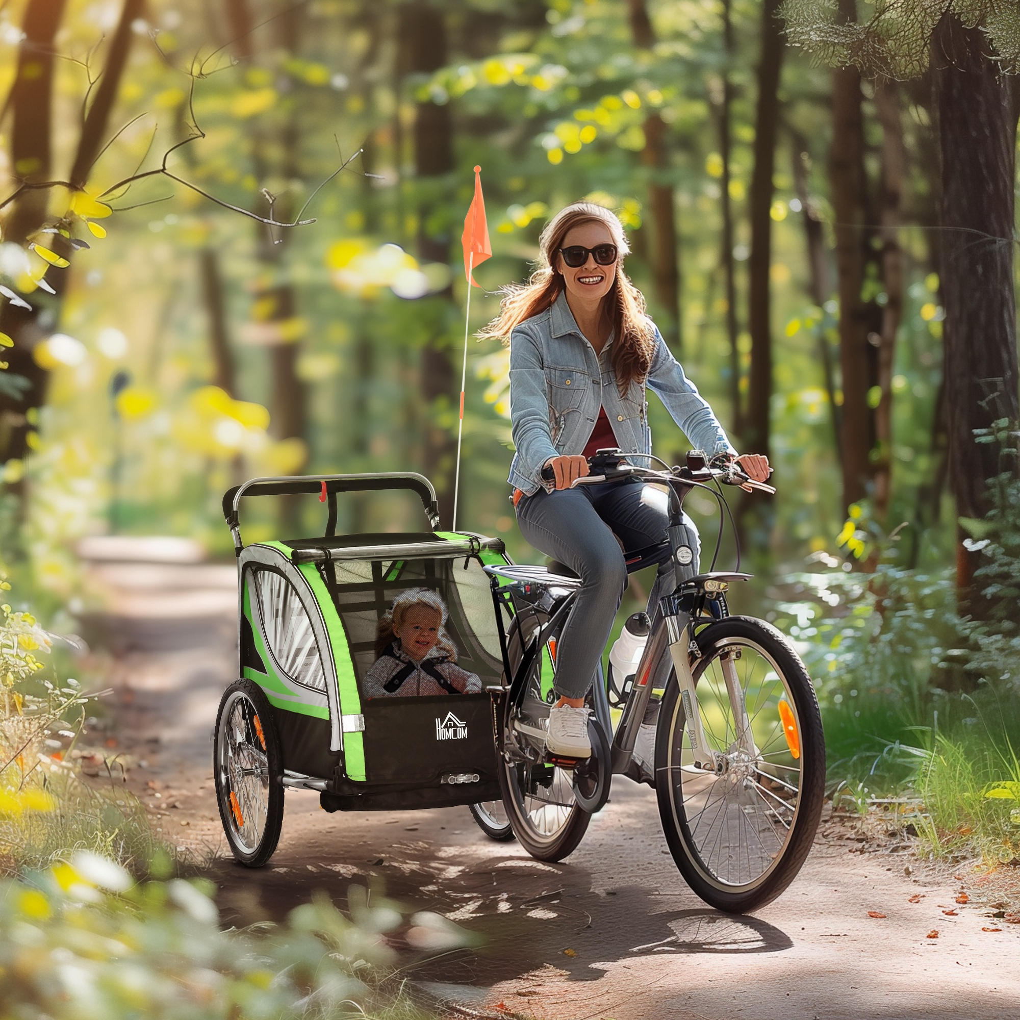 Una donna sorridente con gli occhiali da sole va in bicicletta su un sentiero nel bosco, trainando un rimorchio per bambini.