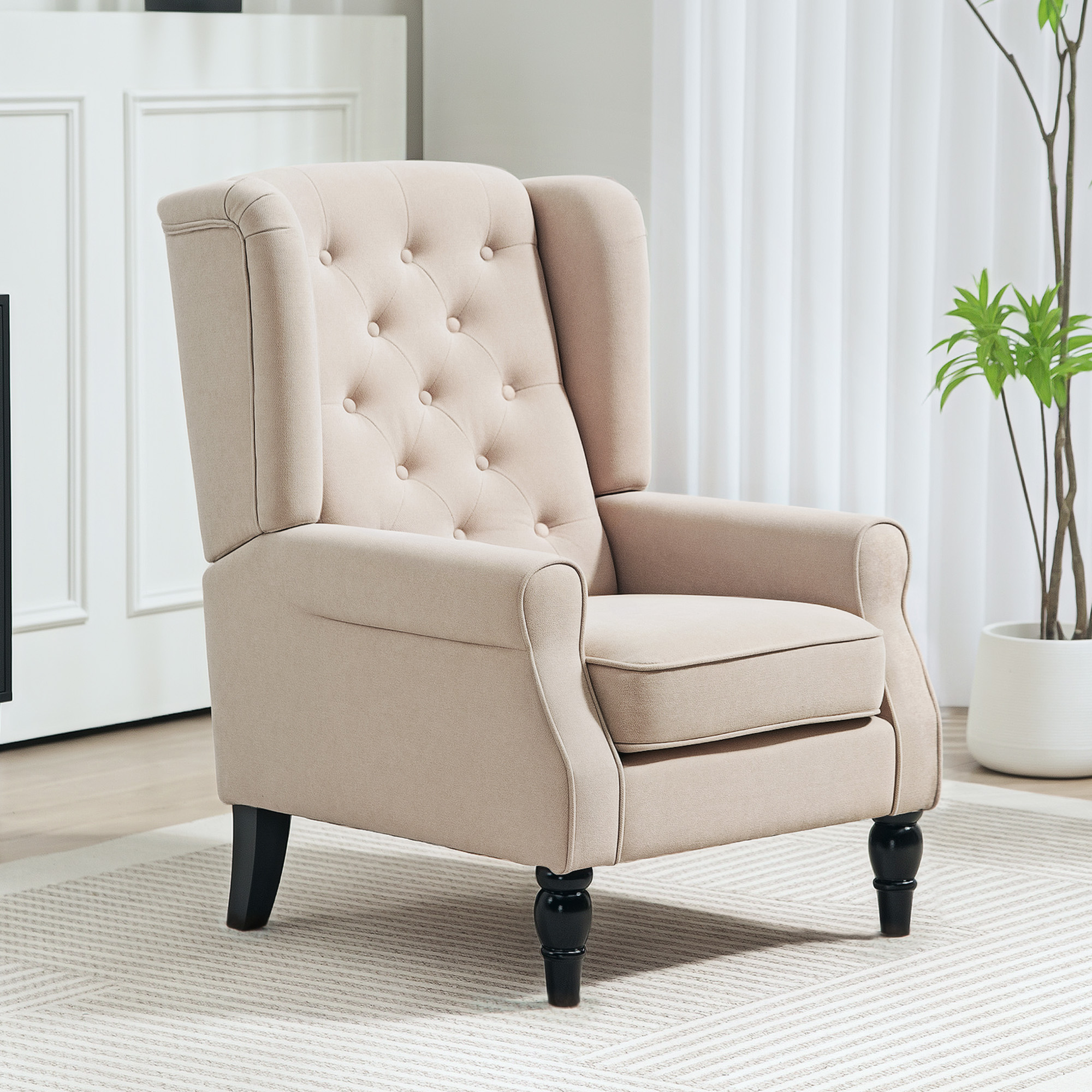 Poltrona orecchiona beige trapuntata con gambe in legno scuro su tappeto bianco e pianta.