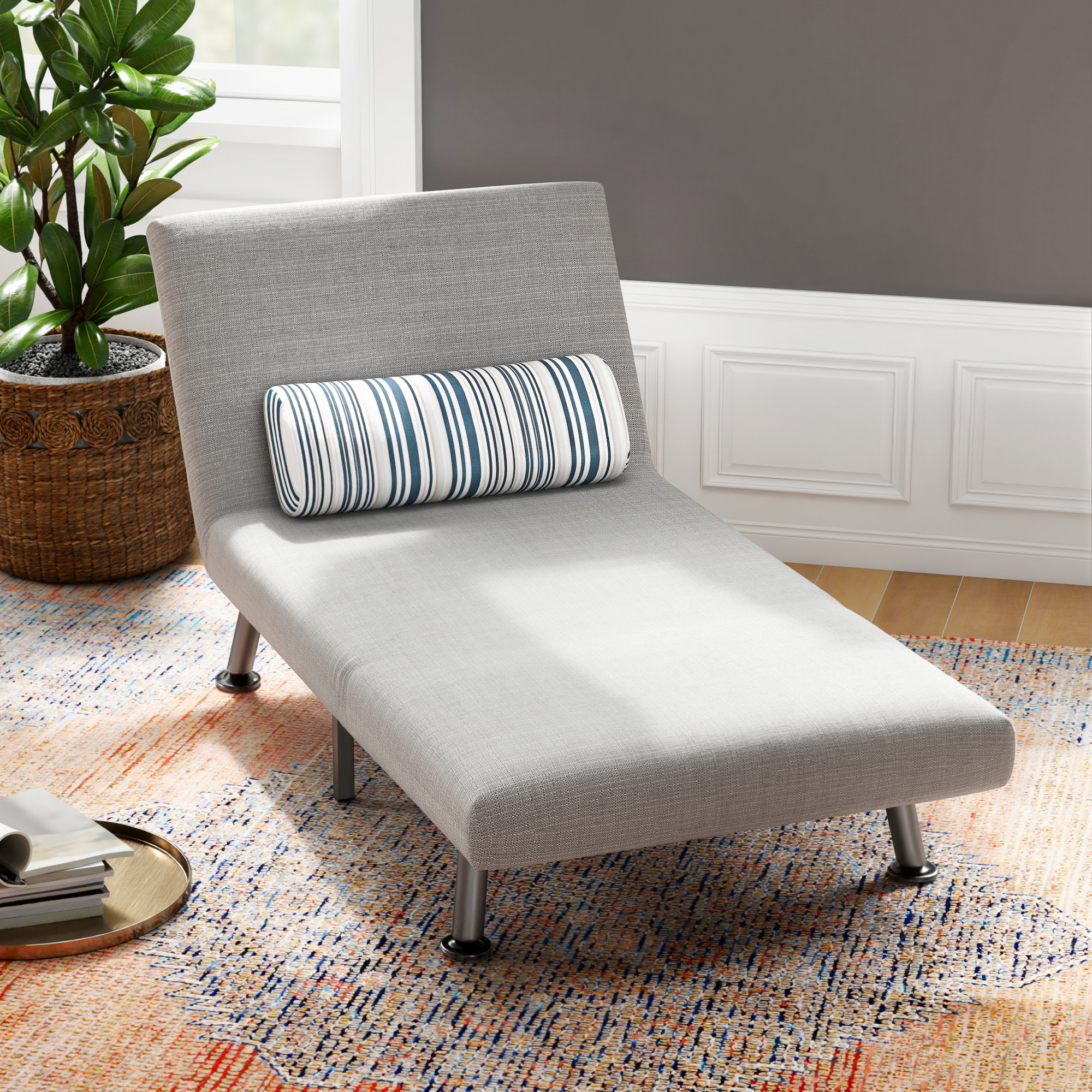 Una chaise longue grigia con cuscino a righe su un tappeto colorato in una stanza con pianta.