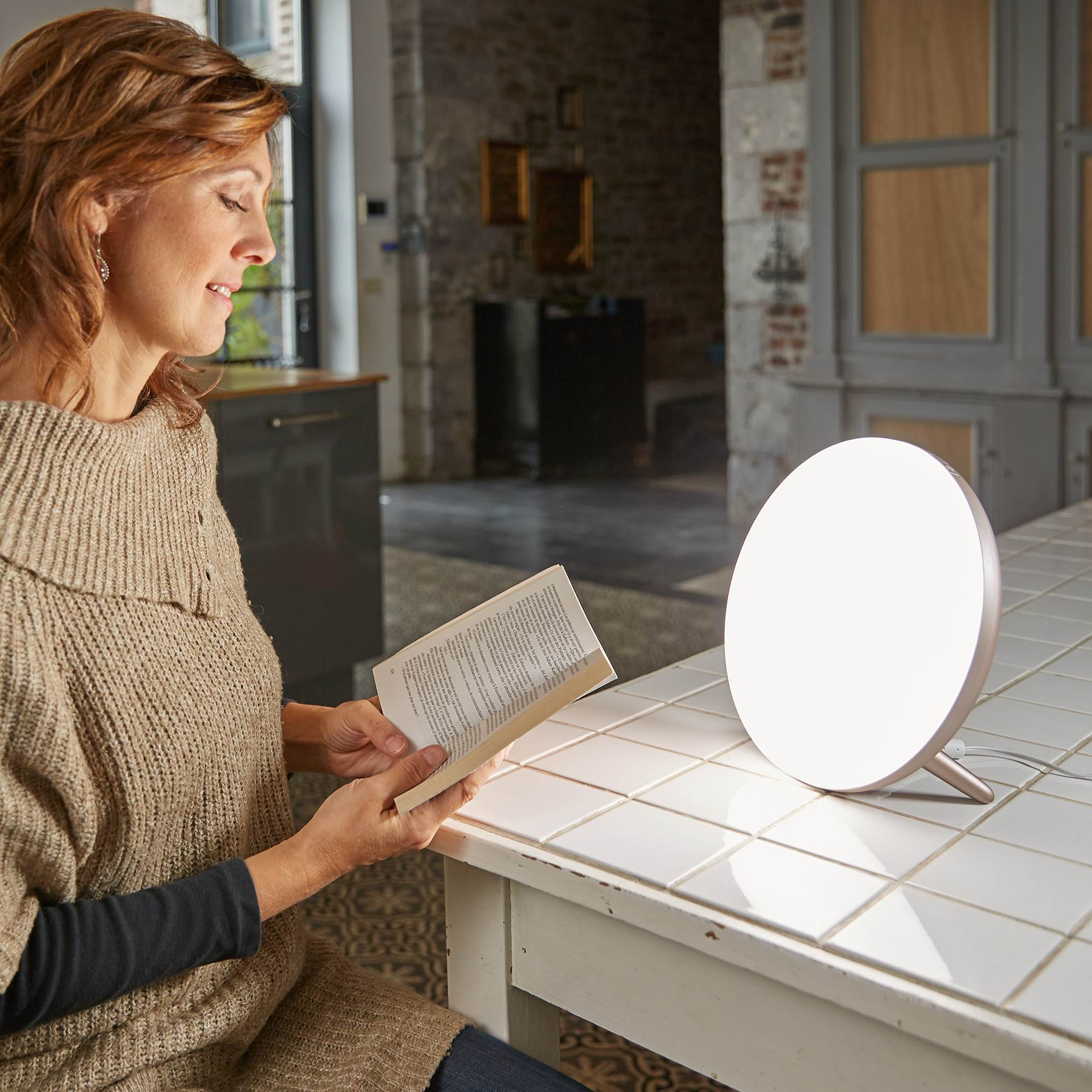 Eine Frau liest ein Buch am Tisch neben einer runden, leuchtenden Lampe.