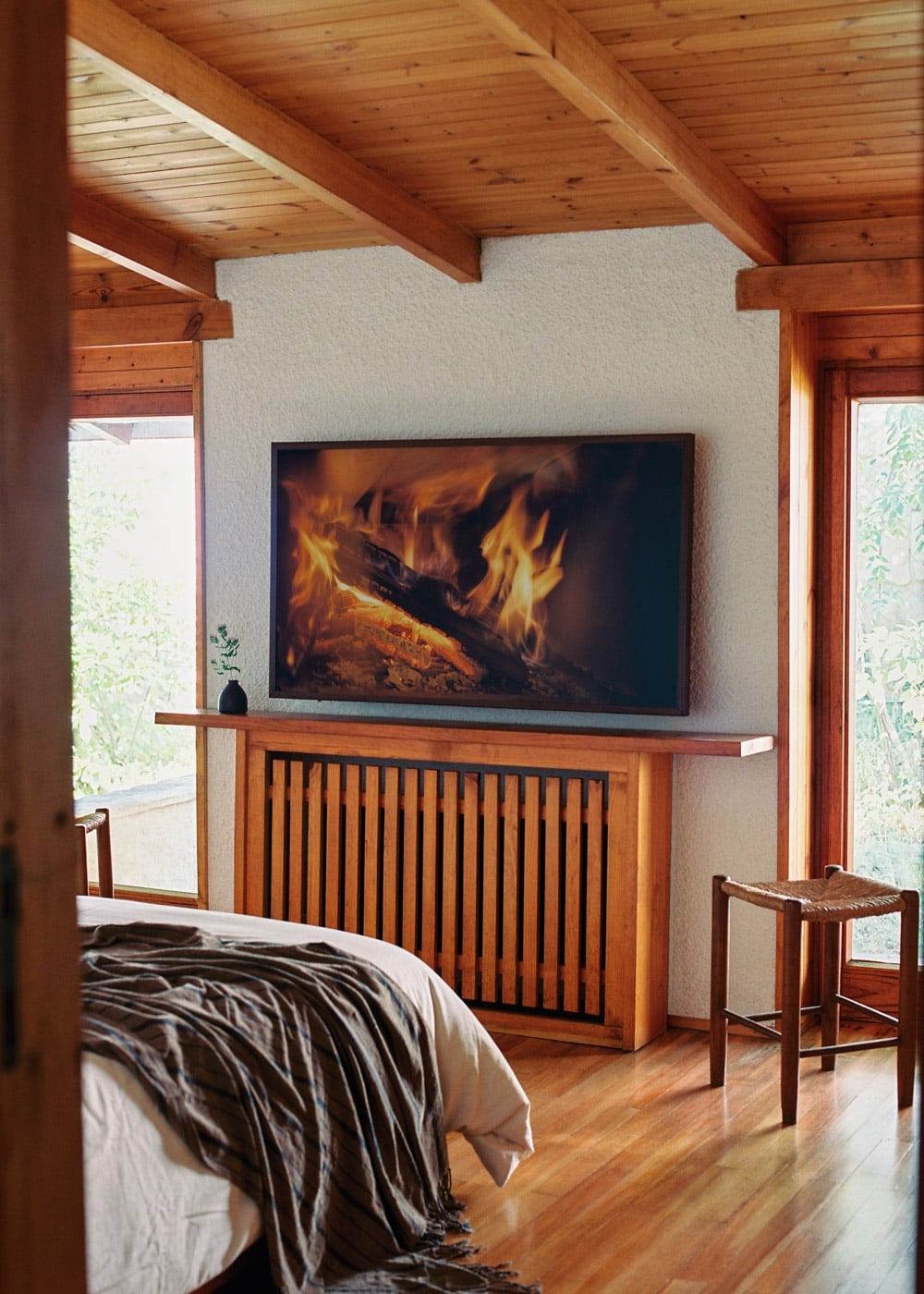 Dormitorio de madera con cama, cubreradiador, imagen de chimenea en pared blanca y pequeño taburete en suelo.
