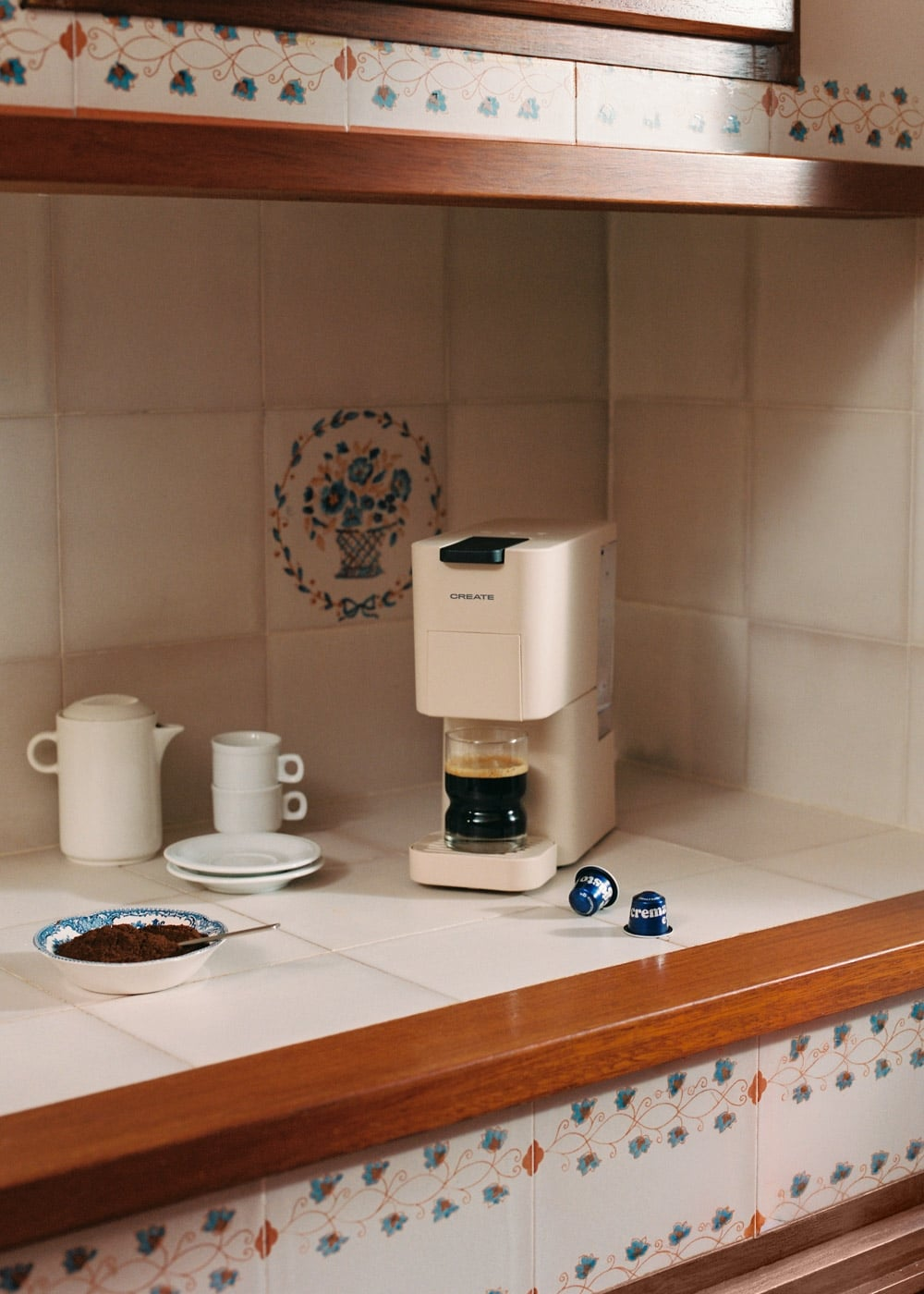 Una cafetera beis sobre azulejos blancos junto a tazas de café y café molido.
