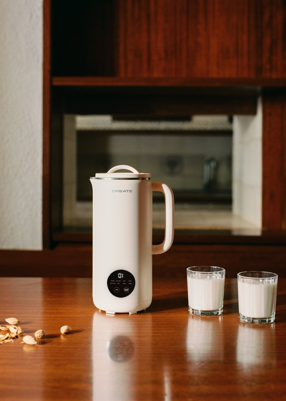 Una máquina de leche vegetal blanca y dos vasos de leche en una mesa de madera, con un mueble de madera de fondo.