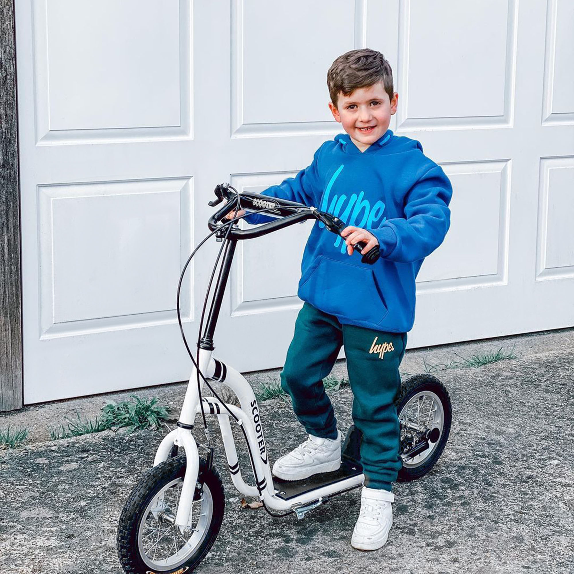 Un ragazzo con felpa blu e pantaloni della tuta verdi si trova su un monopattino bianco davanti a una porta bianca.