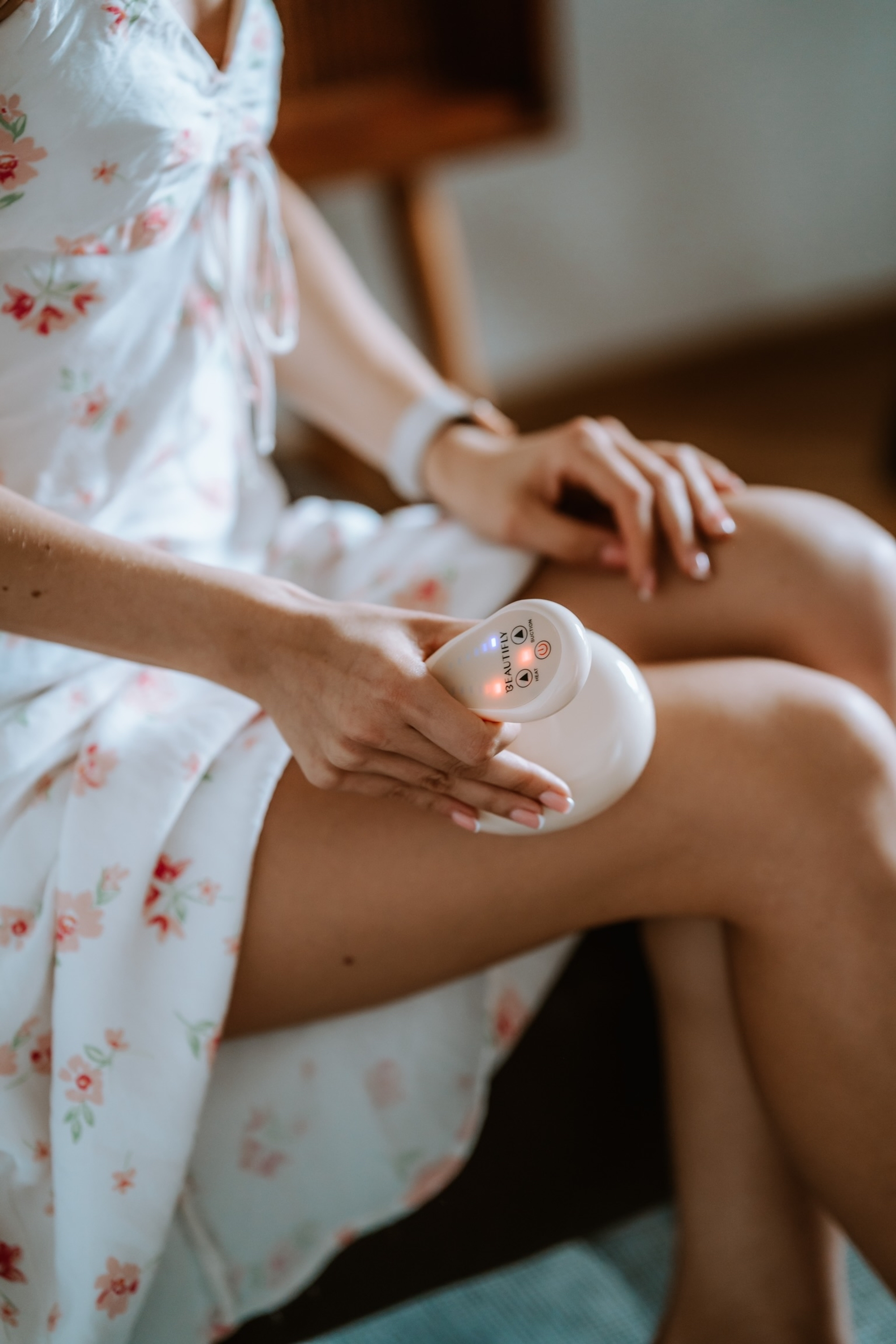 Eine Frau im Blumenkleid hält ein weißes Anti-Cellulite-Gerät auf dem Oberschenkel.
