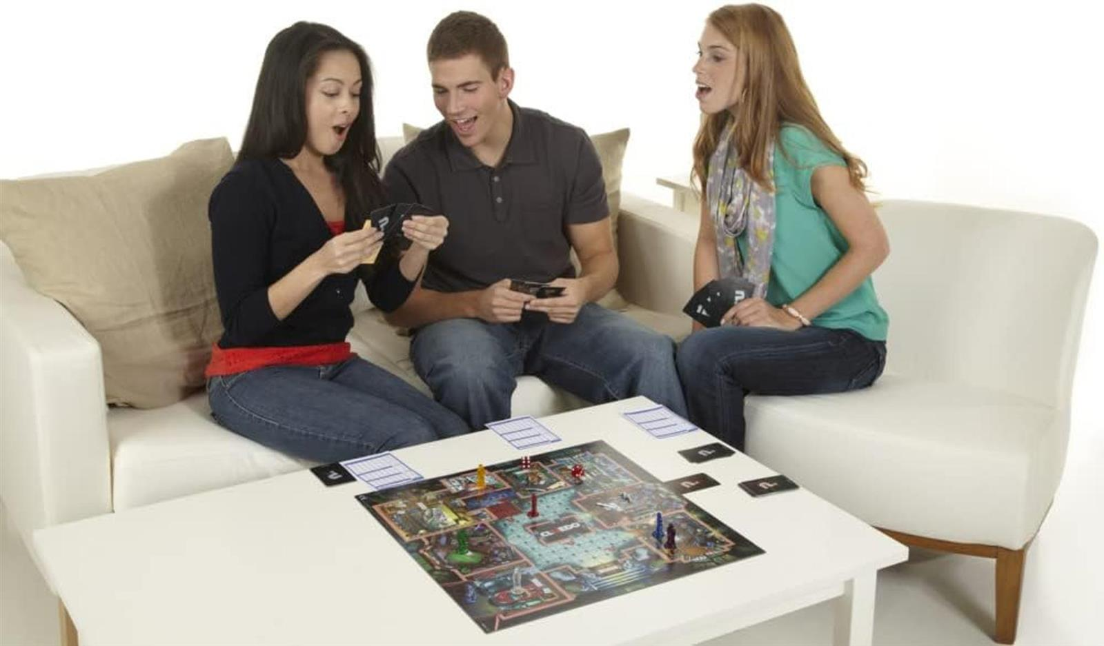 Drei junge Menschen spielen lebhaft ein Brettspiel auf einem weißen Sofa und Tisch.