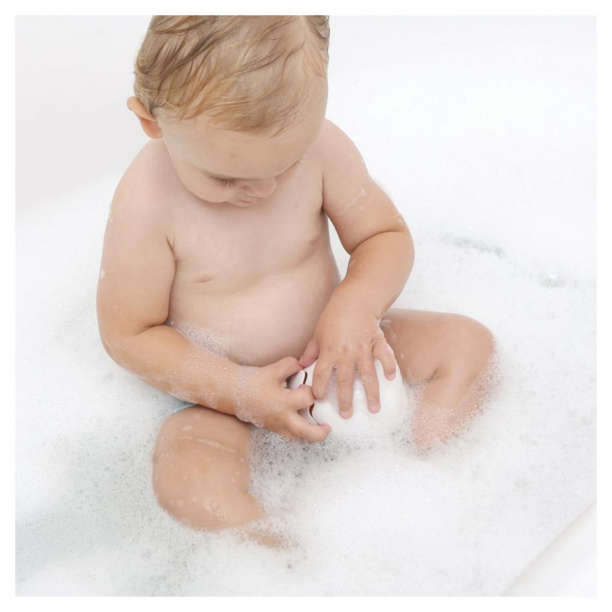 Kleinkind sitzt in einer Badewanne voller Seifenschaum und spielt mit einem weißen Spielzeug.