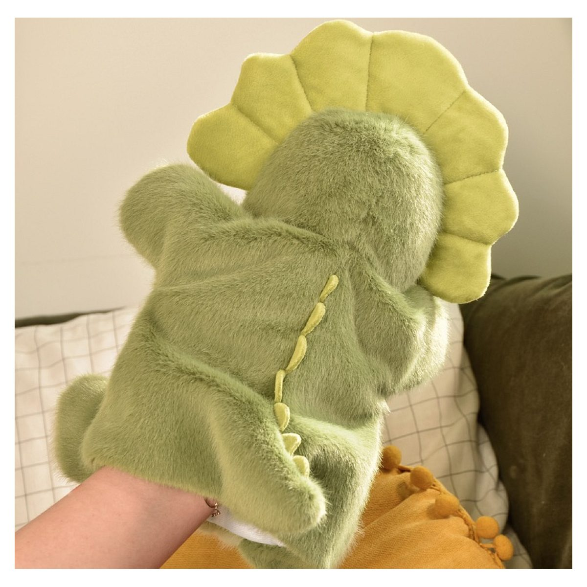 Ein flauschiger grüner Dinosaurier-Handpuppe wird von hinten auf einem Bett gezeigt.