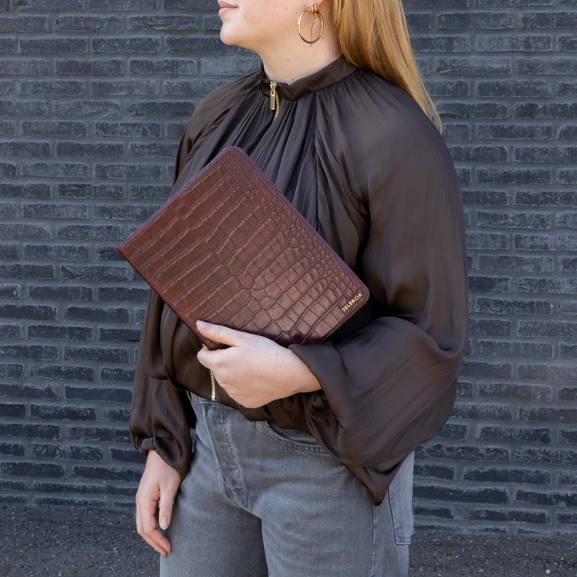 Een vrouw in bruine blouse en grijze jeans houdt een donkerrode mappen met krokodillenstructuur tegen een stenen muur.