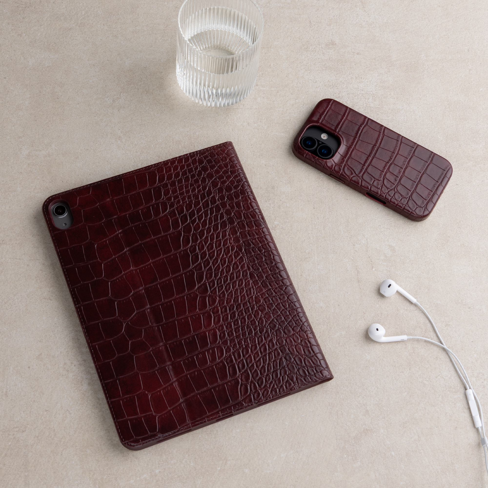 Bordeauxrode krokodillenleren hoesjes voor tablet en smartphone, een glas water en witte oordopjes op een beige oppervlak.