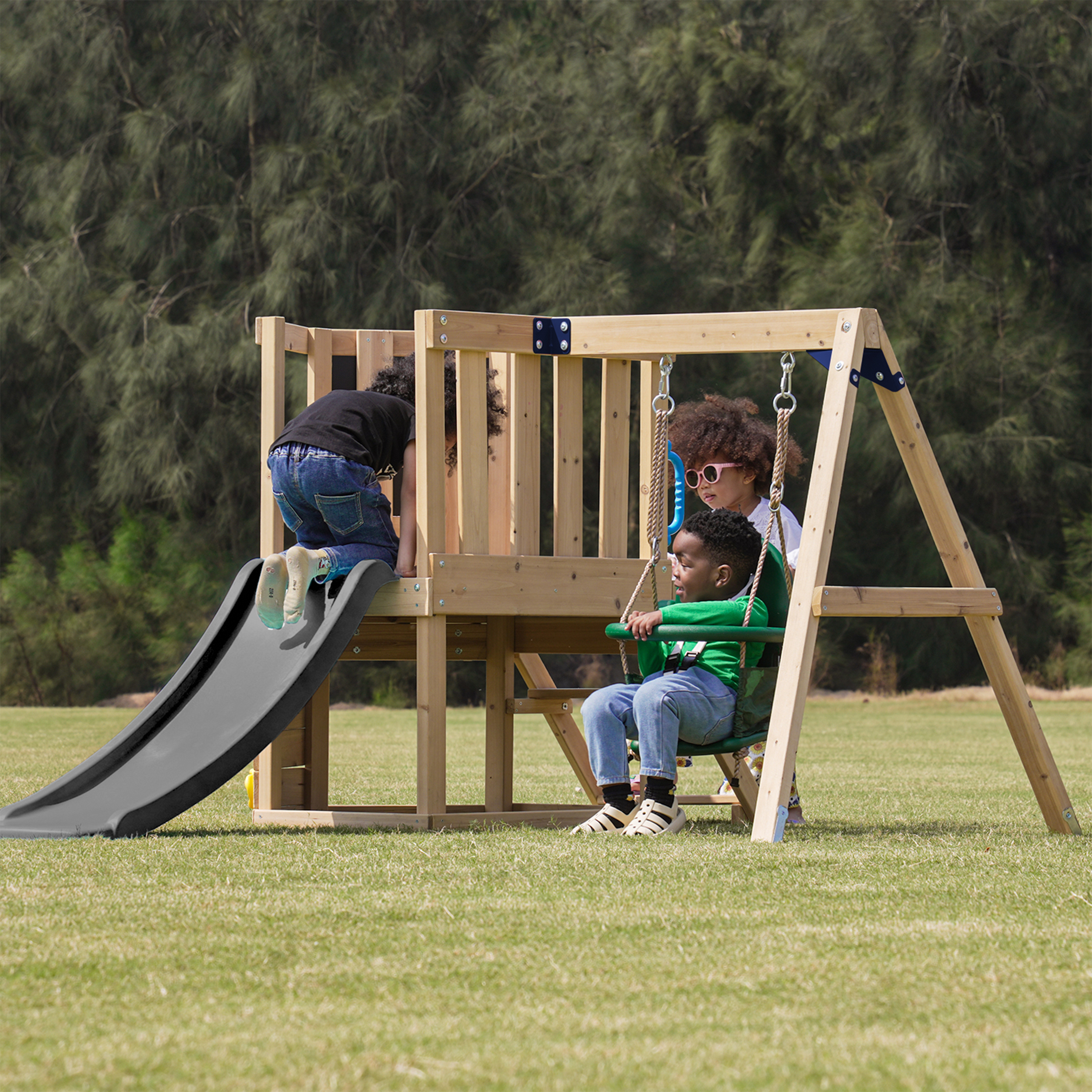 Drei Kinder spielen auf einem Holzd Spielplatz auf Rasen, eines auf einer Rutsche, zwei auf einer Schaukel.