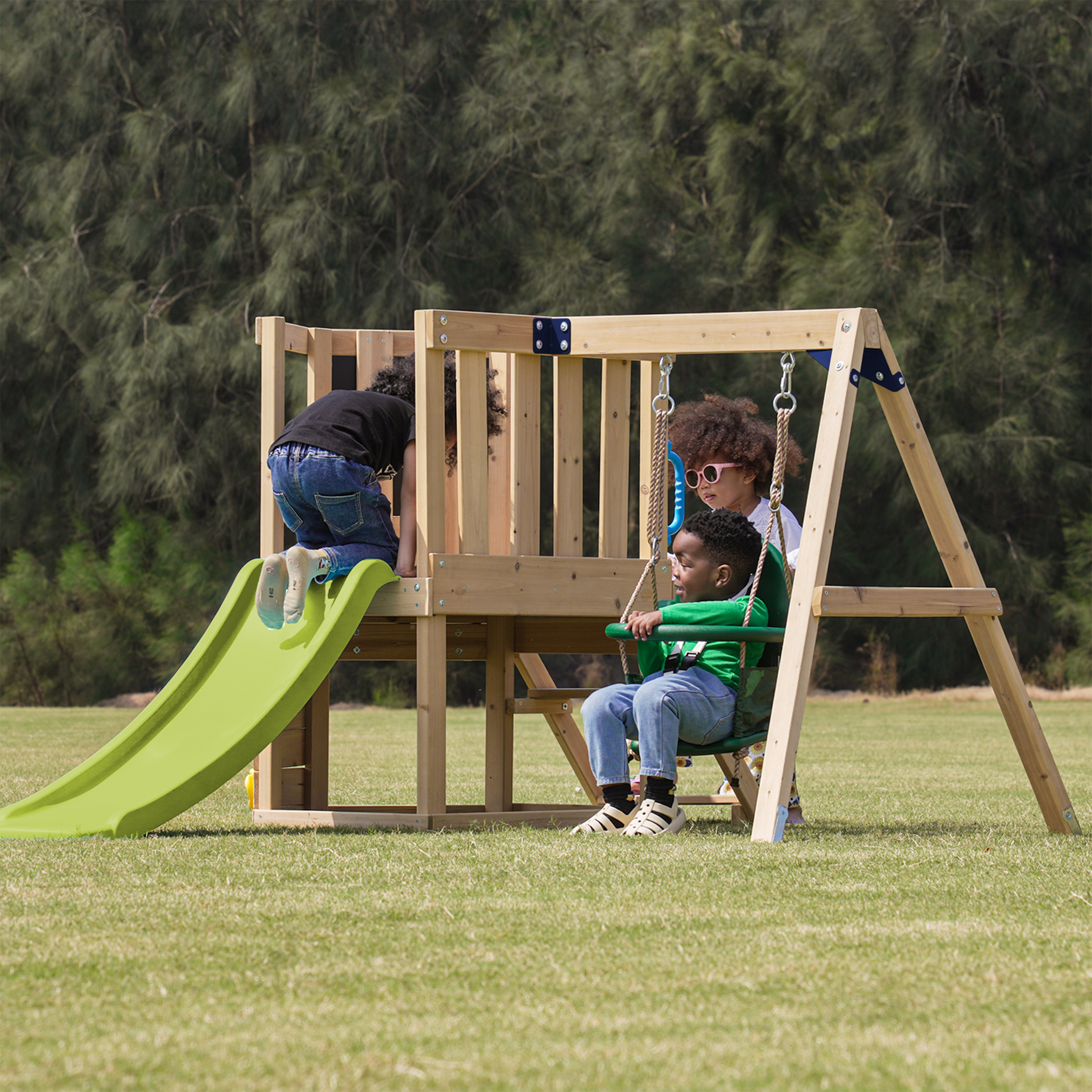 Drei Kinder spielen auf einem Holzspielplatz mit grüner Rutsche und Schaukeln auf einer Wiese mit Bäumen.