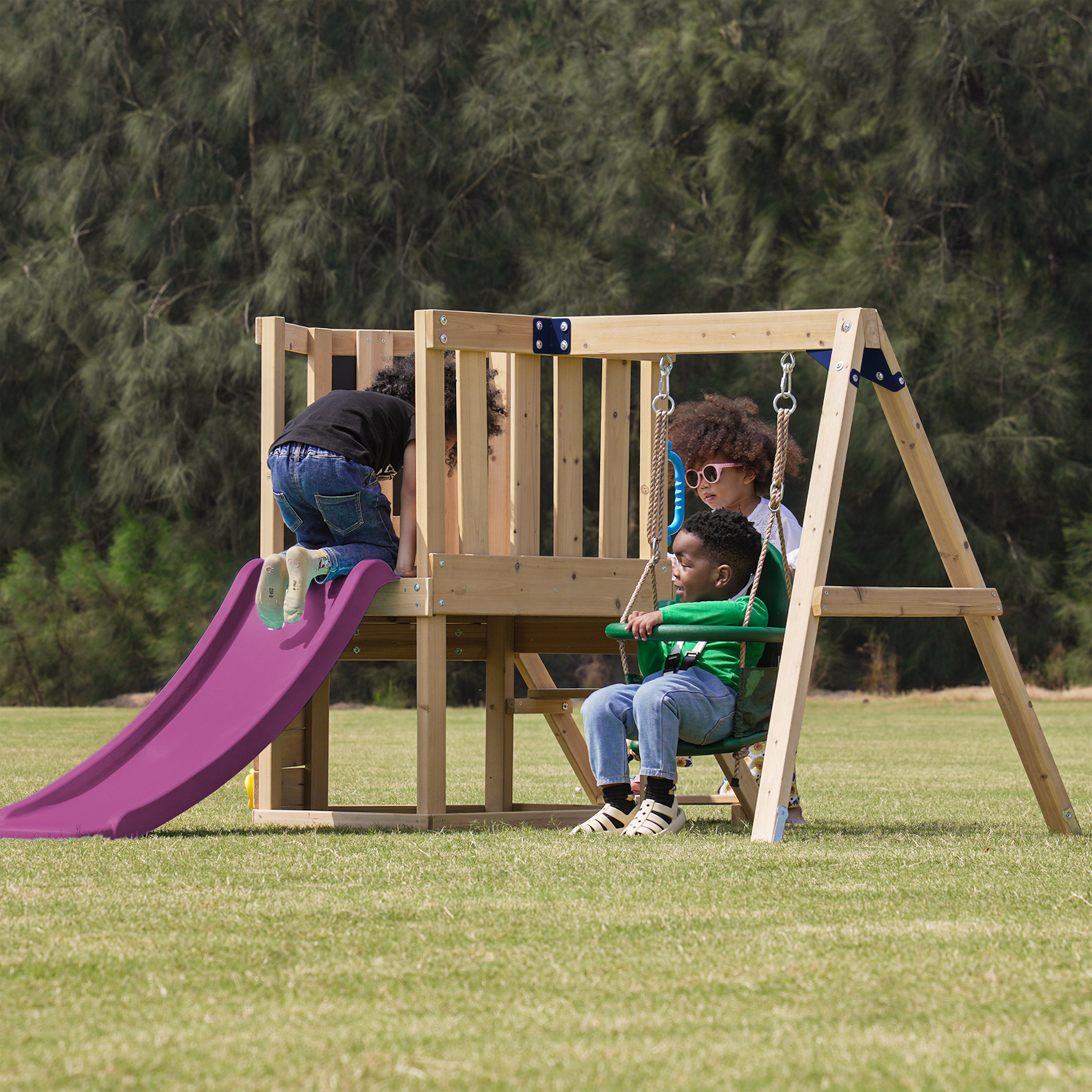 Drei Kinder spielen auf einem Holzspielplatz mit Schaukel und Rutsche im Freien.