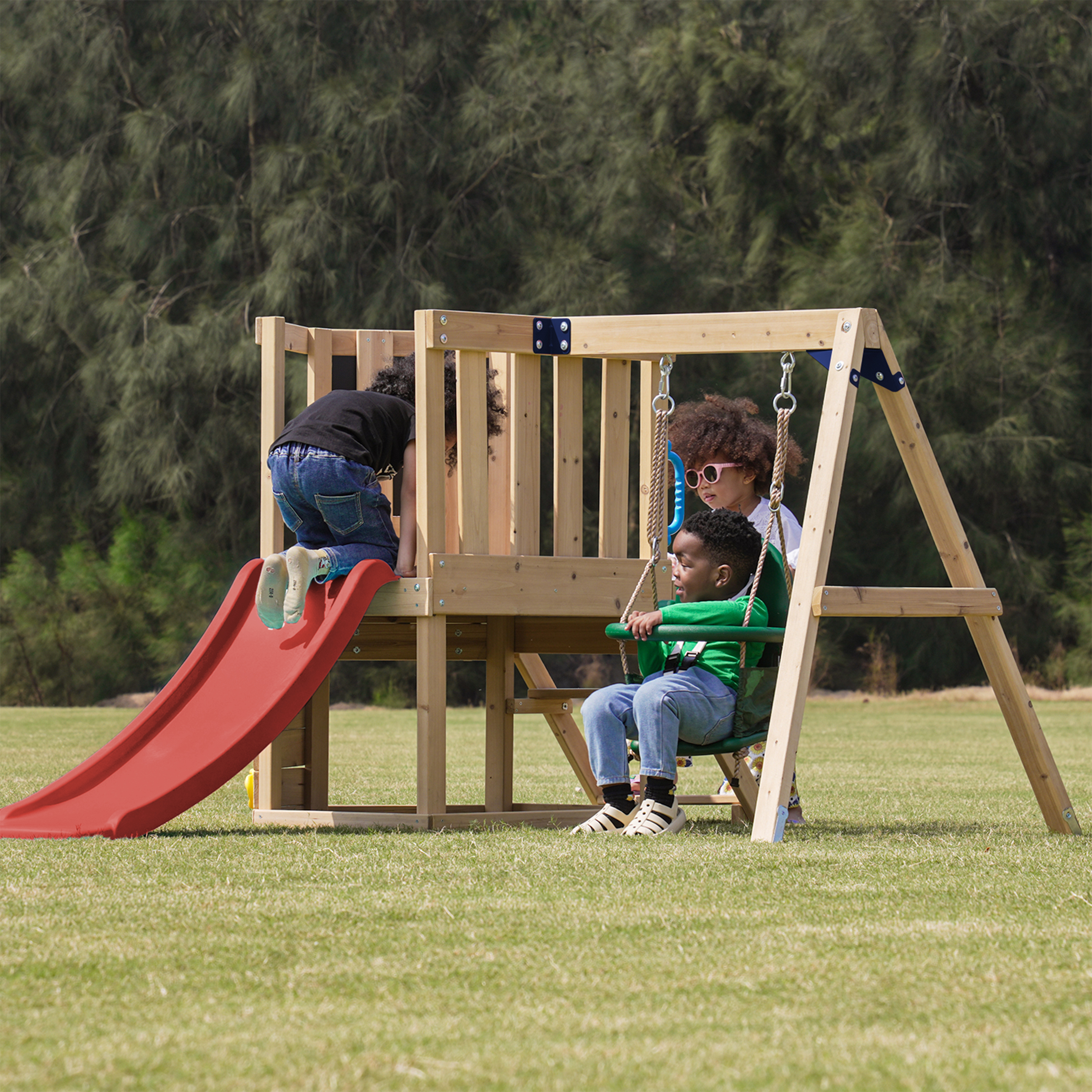 Drei Kinder spielen auf einem Holzspielplatz mit roter Rutsche und Schaukeln auf einer Wiese.