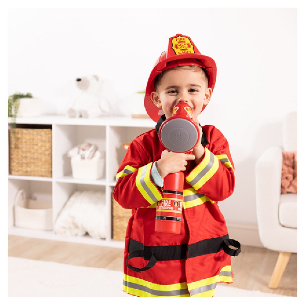 Niño pequeño disfrazado de bombero sostiene un extintor de juguete rojo en una habitación infantil.