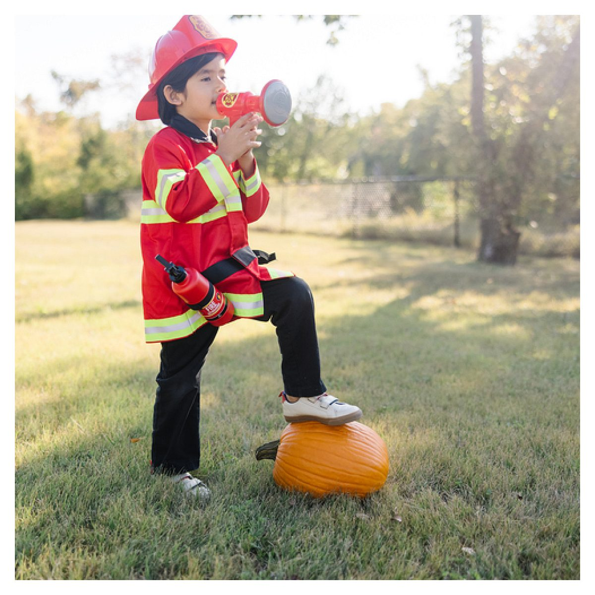 Un niño disfrazado de bombero habla por un megáfono con un pie en una calabaza sobre el césped.