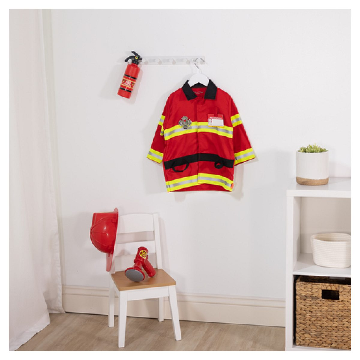 Un disfraz de bombero rojo cuelga en la pared, abajo hay una silla con casco y megáfono, al lado de un estante blanco.