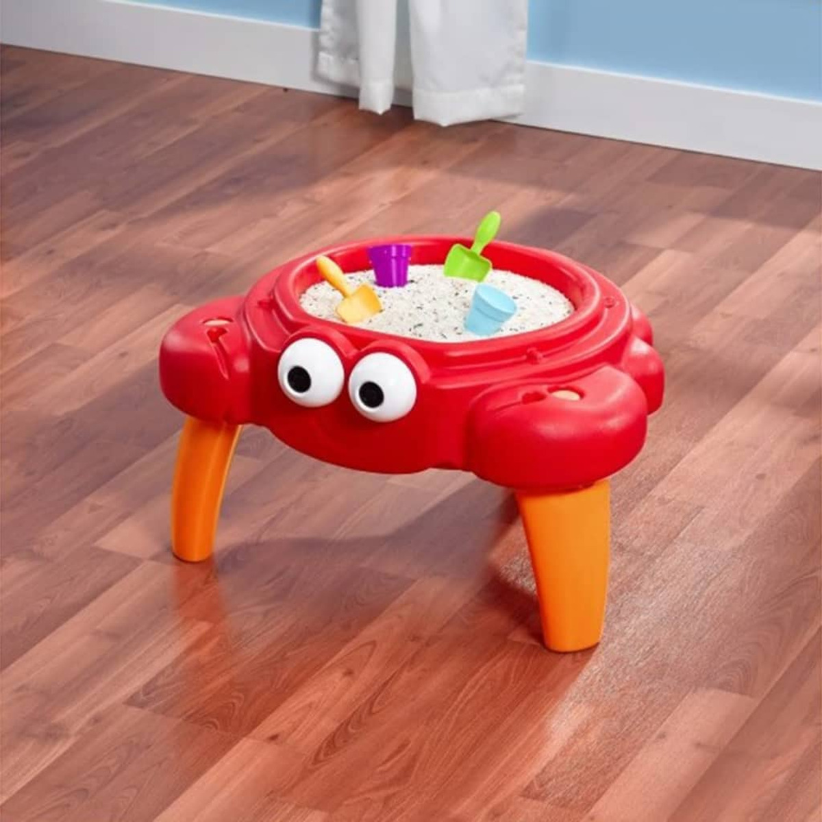 Ein roter krebssicherer Sandtisch mit weißem Sand und buntem Spielzeug auf Holzboden.
